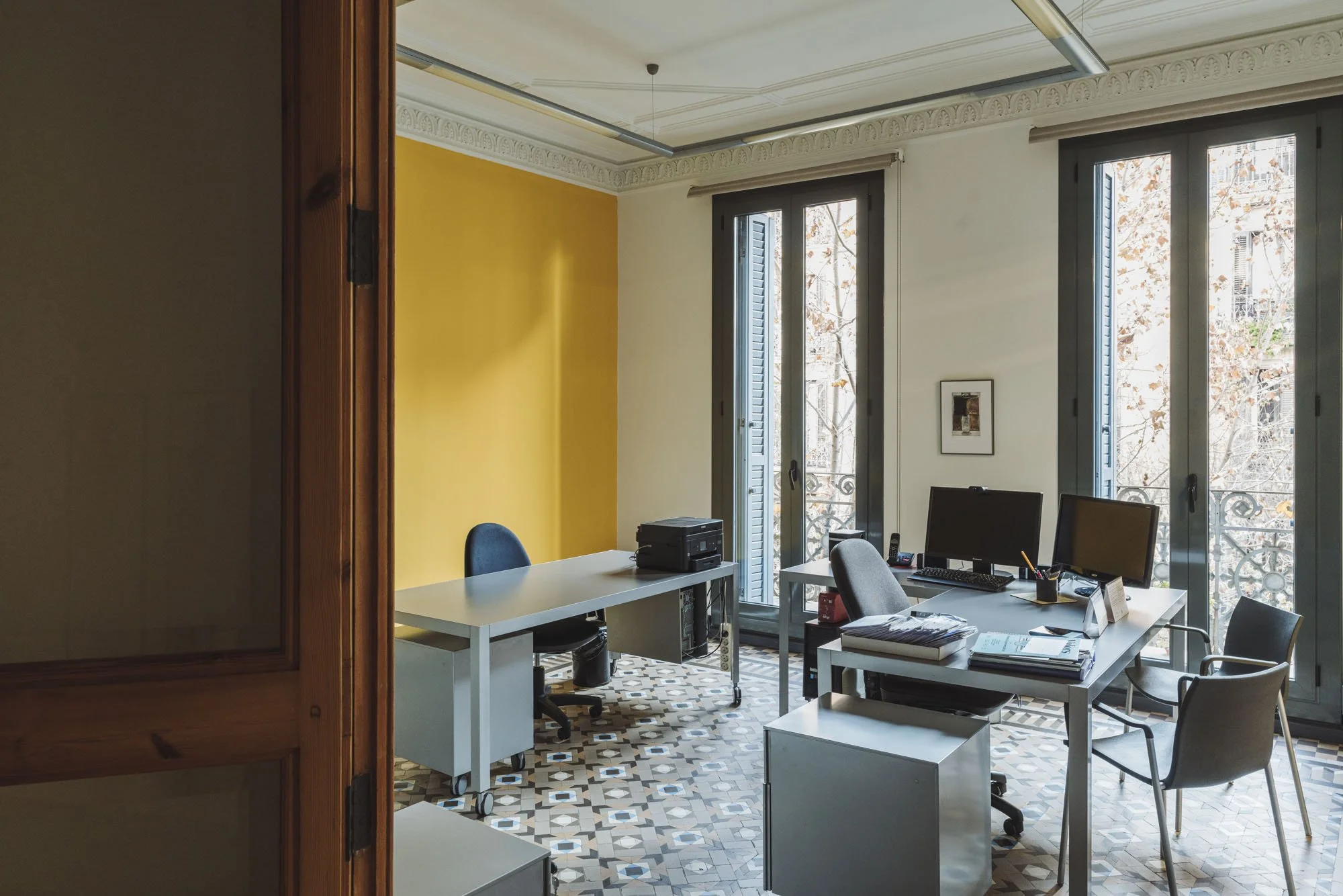 Oficina con dos escritorios, con varias sillas, computadoras, documentos y un teléfono, con ventanas grandes y puertas de balcón, paredes blancas y un muro amarillo, con piso de azulejos decorativos.