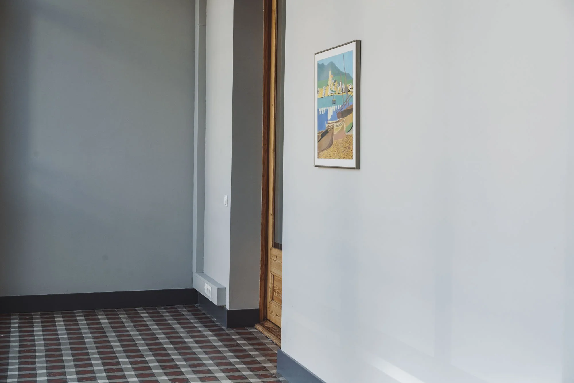 Paredes blancas en un interior de casa, con un cuadro decorativo en la pared y un piso de baldosas con patrón de cuadros en tonos rojo y gris.