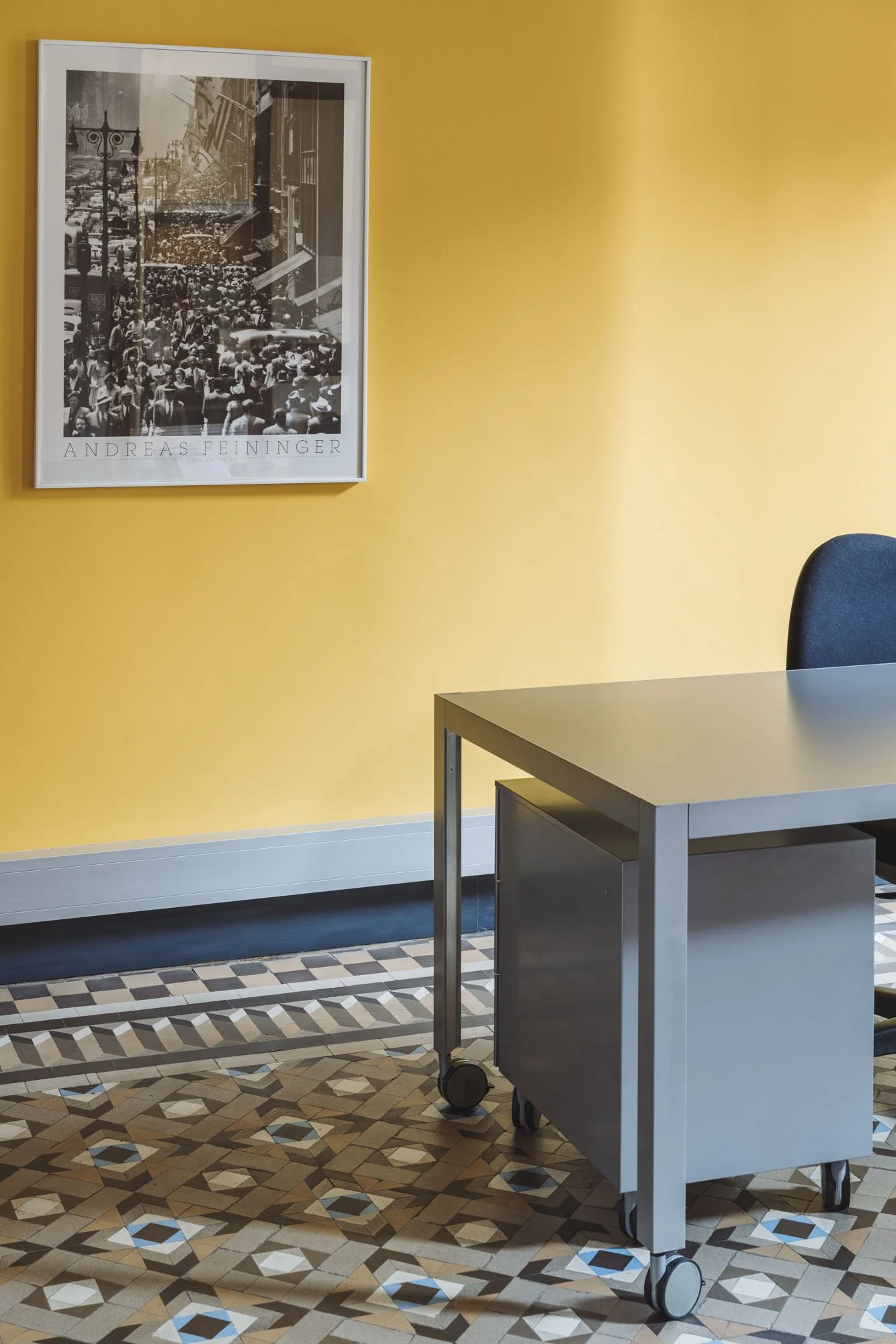 Pared amarilla con cuadro de fotografía y texto, escritorio gris, silla azul y piso con mosaico de patrones geométricos en tonos tierra, azul y blanco.