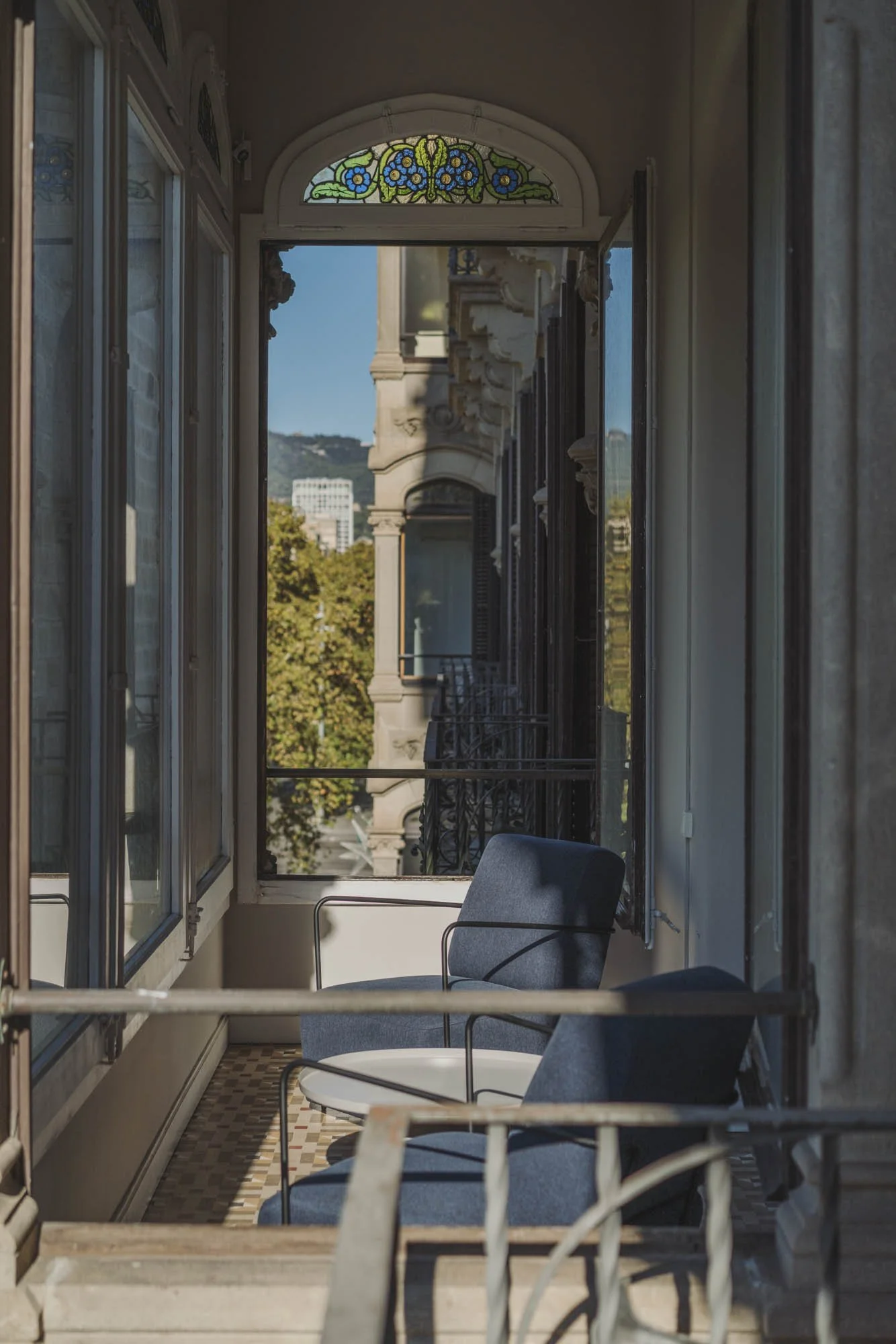 Balcón con sillones y vista a edificios y árboles, con ventanales y decoración de vidrieras en un entorno urbano.