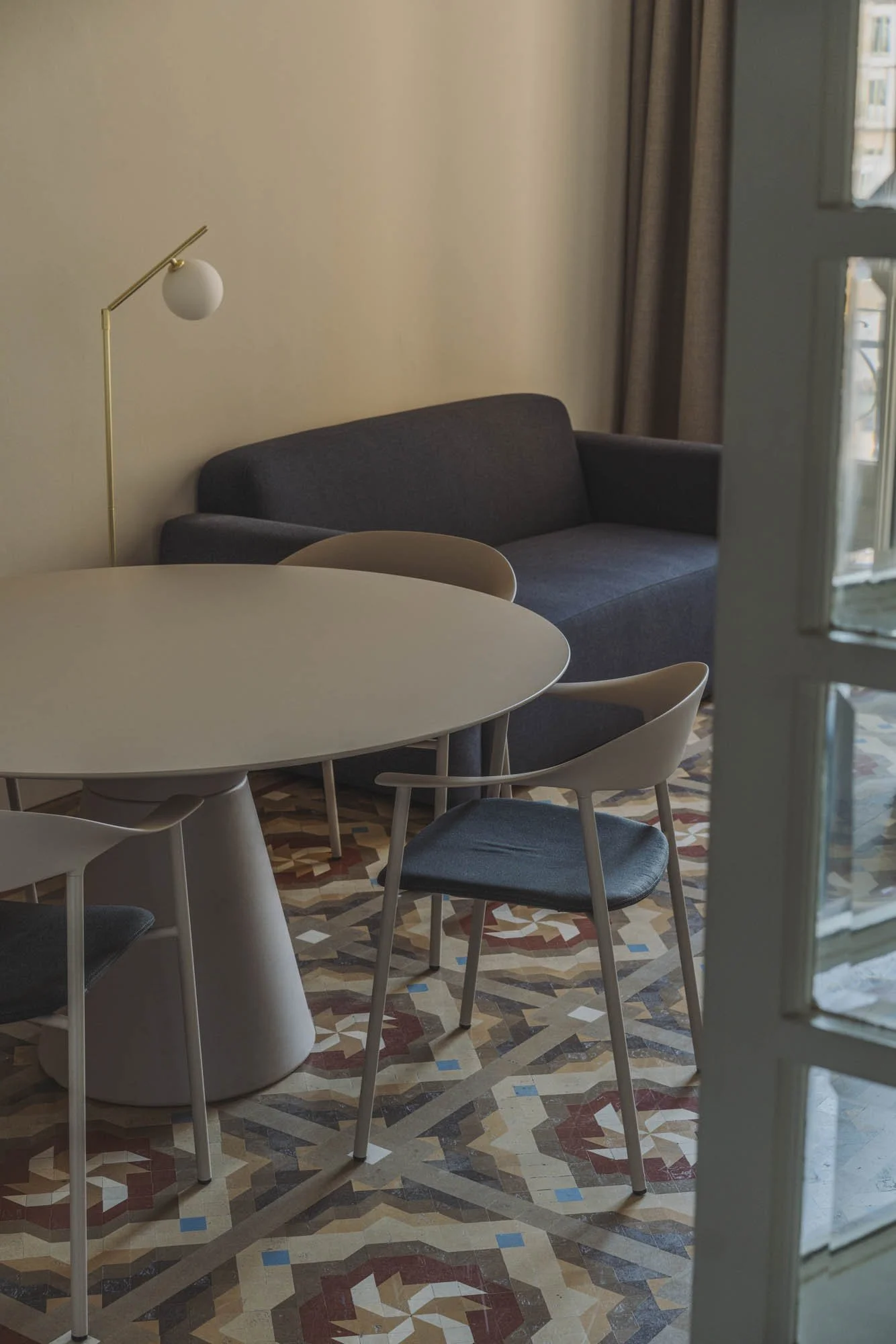 Sala de estar con sofá gris, mesa redonda beige, sillas con asientos grises, suelo con azulejos decorativos y pared beige, con ventana y cortinas beige.