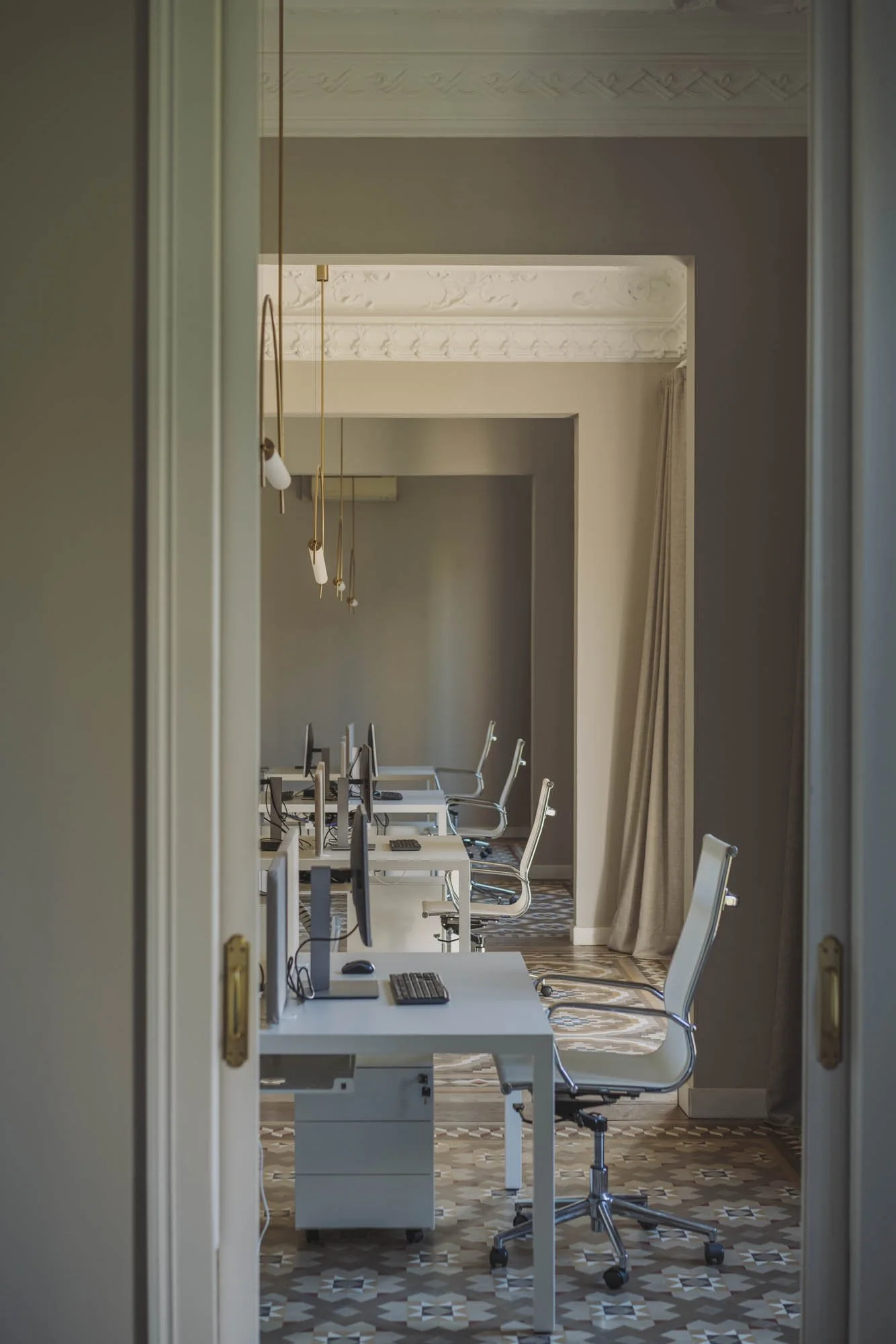 Sala de oficina con varias estaciones de trabajo y sillas blancas, vista a través de una puerta. Muebles blancos, ordenadores y cortinas beige en el fondo, con un suelo de patrón geométrico.