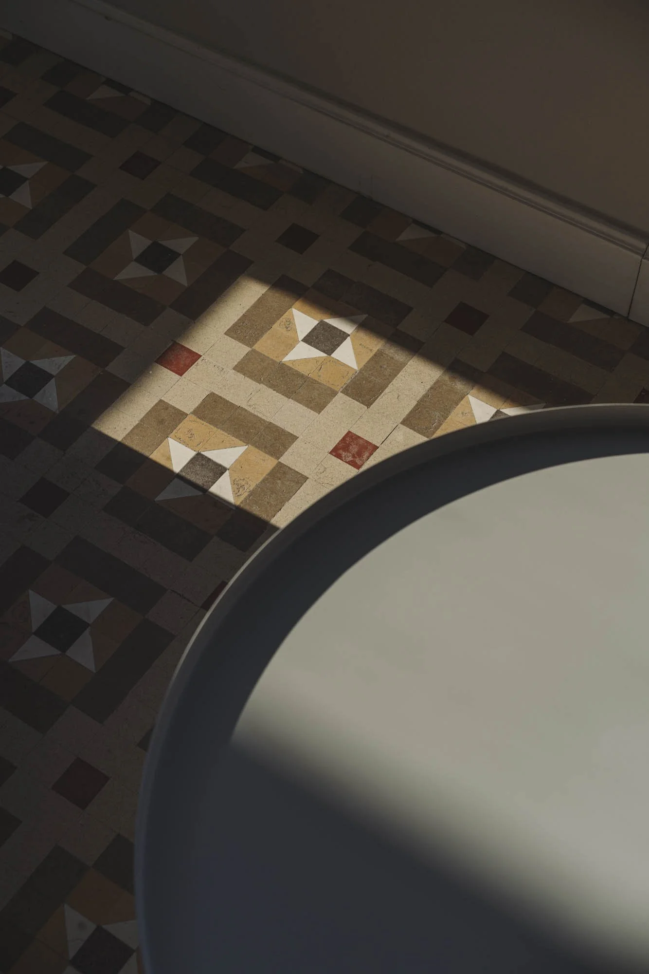 Vista de un piso con baldosas decorativas en tonos marrones, beige, y rojo, con un patrón geométrico, parcialmente cubierto por la sombra de una ventana y una mesa redonda de color claro en la esquina inferior derecha.