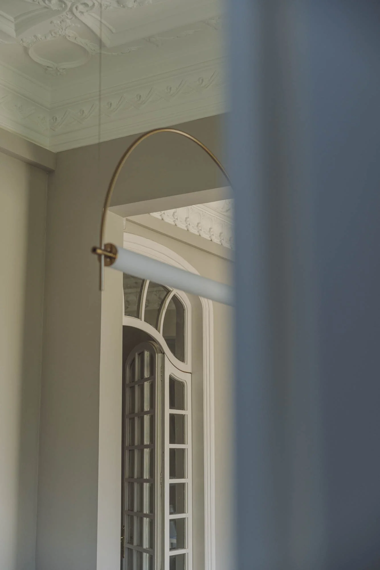 Vista de una ventana con arco y varias ventanas pequeñas, enmarcado por un muro en un ambiente interior con molduras decorativas en el techo y una estructura de lámpara moderna colgante