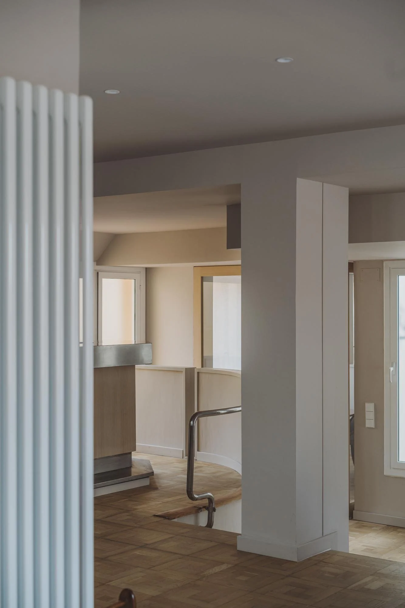 Interior de una casa con paredes blancas, ventanas con marco de madera, piso de madera y una barandilla de metal en una escalera.