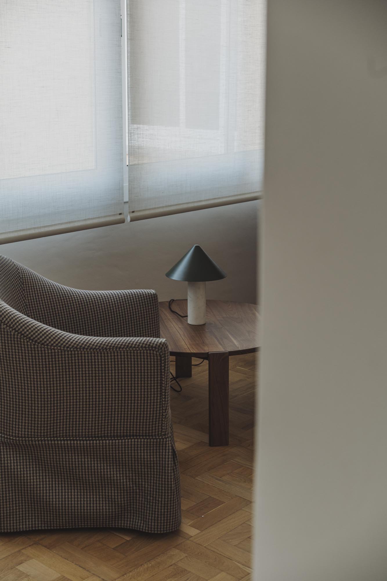 Una sala con sillones de tela con patrón de cuadros pequeños y una mesa de madera con una lámpara moderna de base cilíndrica y pantalla cónica negra. Fondo con ventanas cubiertas con cortinas blanco traslúcidas.