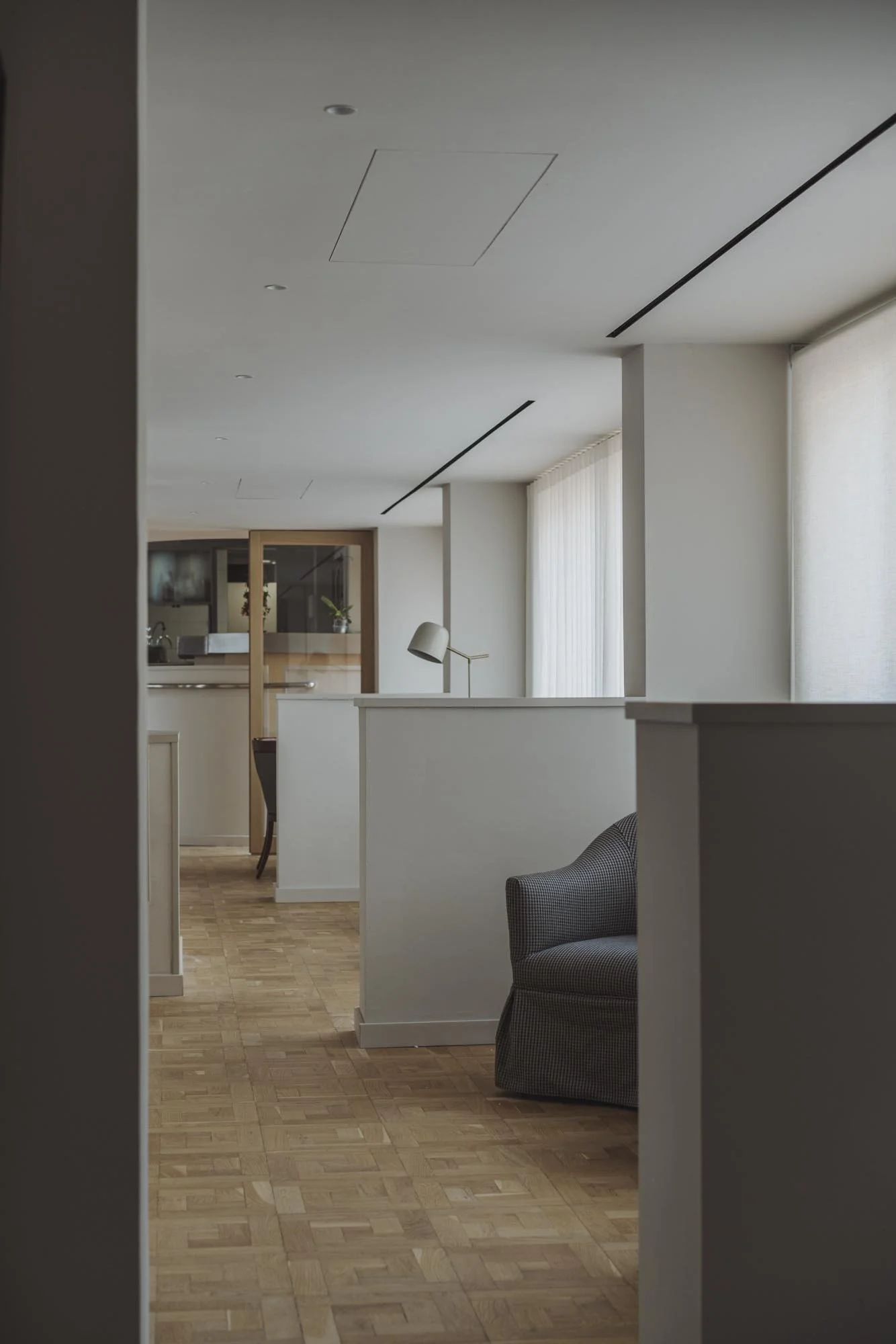 Interior de una sala con paredes blancas, piso de madera clara, cortinas blancas y un mueble con lámpara de escritorio, con una zona de estar con un sillón gris y una mesa en el fondo.