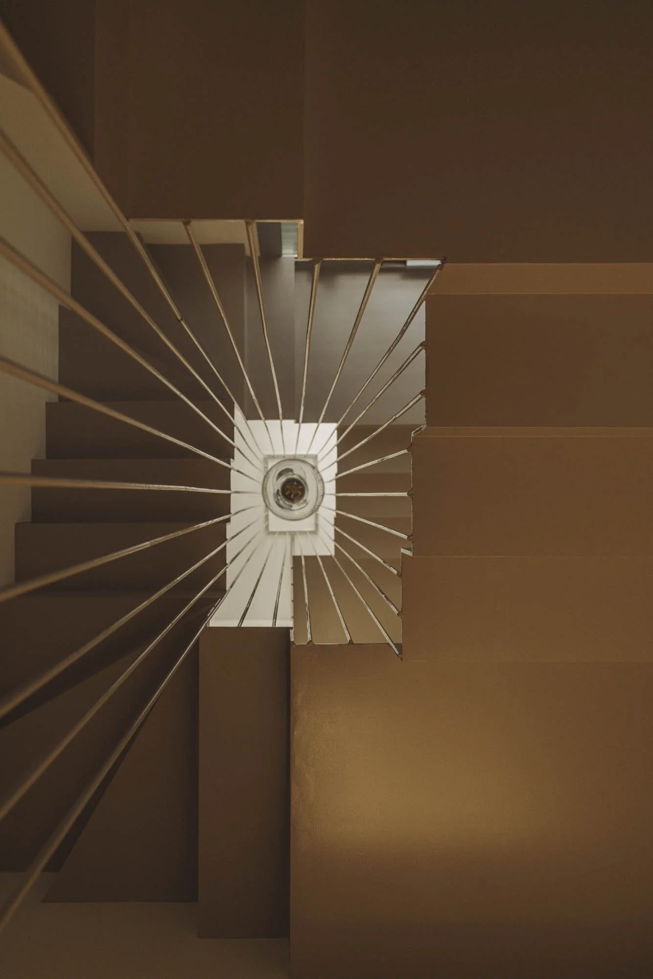 Vista desde arriba de una escalera de color marrón con barandilla de barrotes metálicos, enfocando hacia la luz en la parte superior del descansillo.