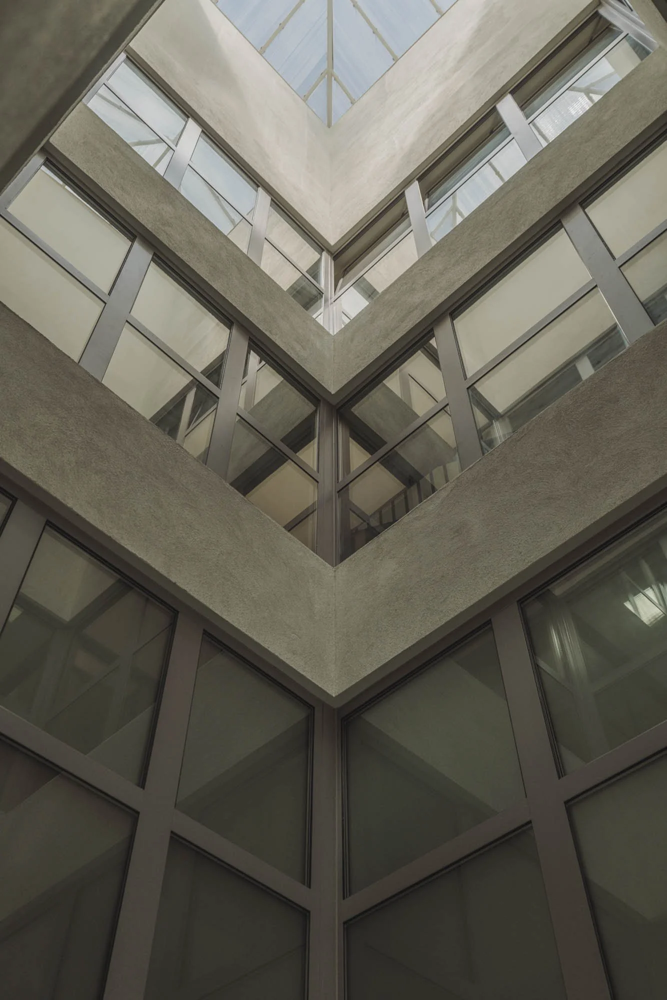 Vista desde abajo hacia el interior de un edificio con paredes de cristal y concreto, mostrando varios niveles con ventanas de vidrio y un techo de vidrio en forma de pirámide.