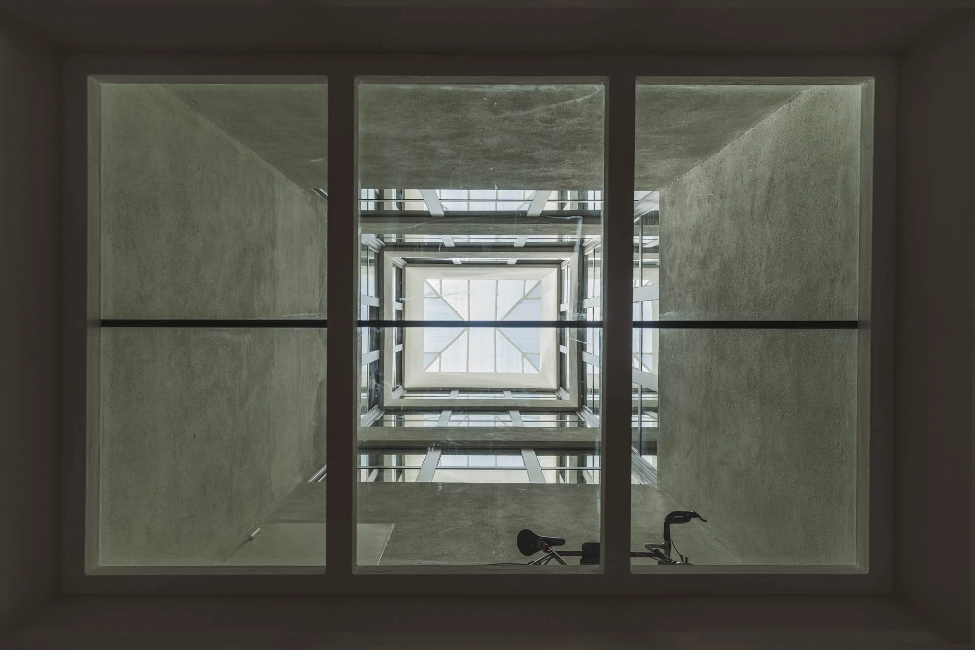 Vista desde abajo de un patio de un edificio con paredes de concreto y un cuadro de escaleras que se extiende hacia arriba, con un ciclista en la esquina inferior derecha.