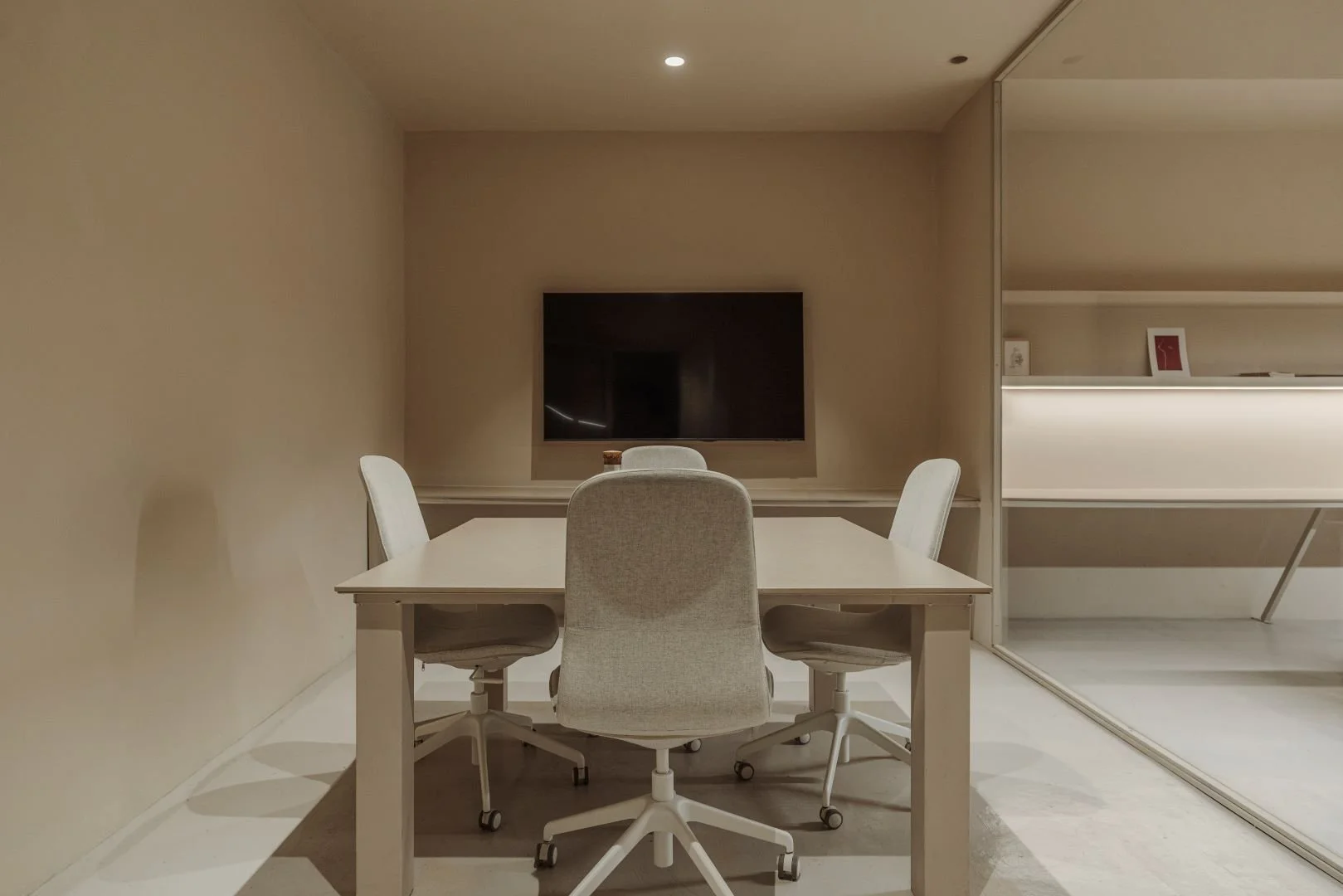 Sala de reuniones con mesa blanca y sillas ergonómicas, pantalla en la pared y una habitación secreta con estanterías visible a la derecha.