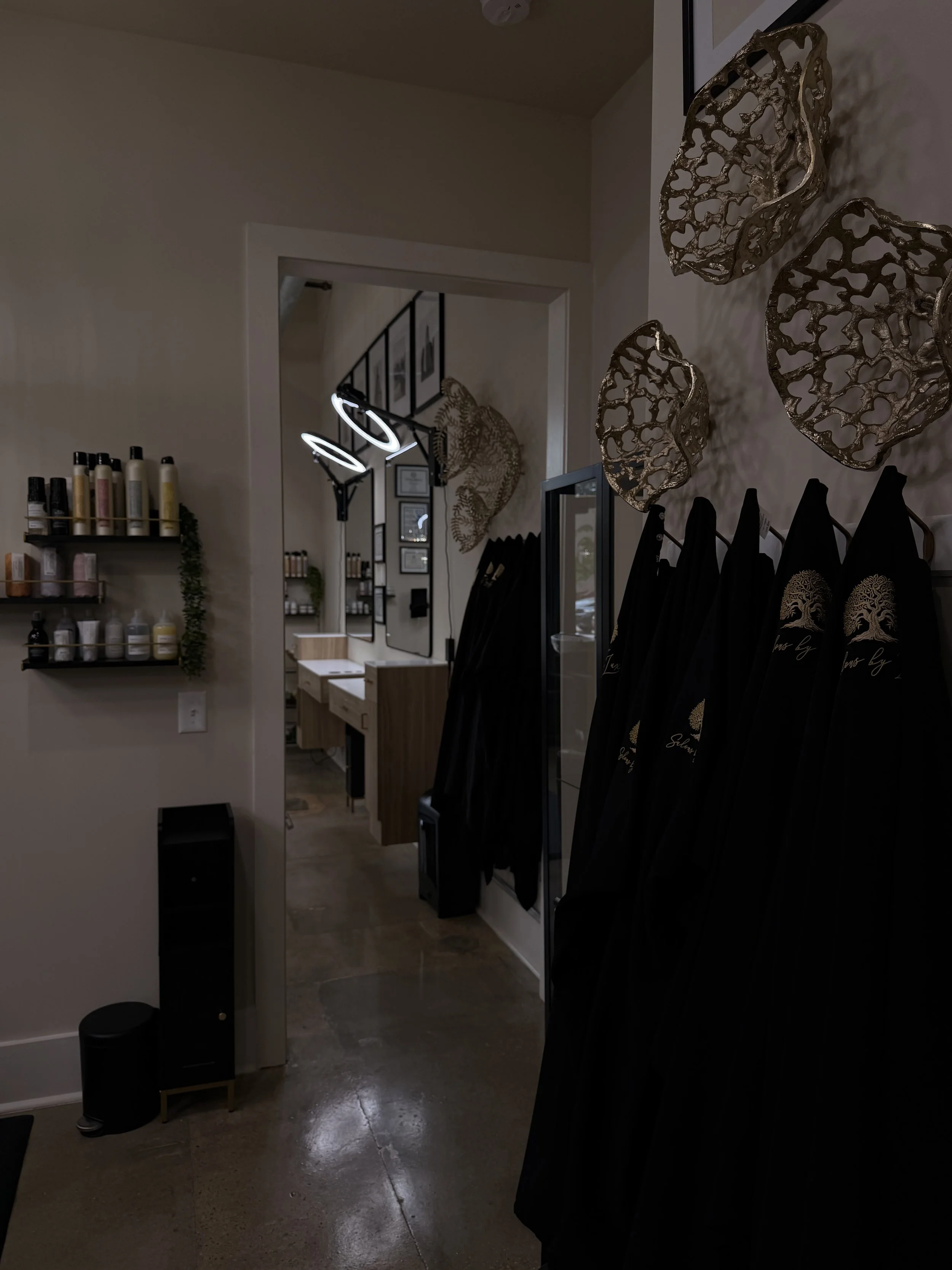 Interior of a salon or boutique featuring black robes hanging on hooks, decorative wall sculptures, a wall-mounted shelf with bottles, and a neon sign in the background.