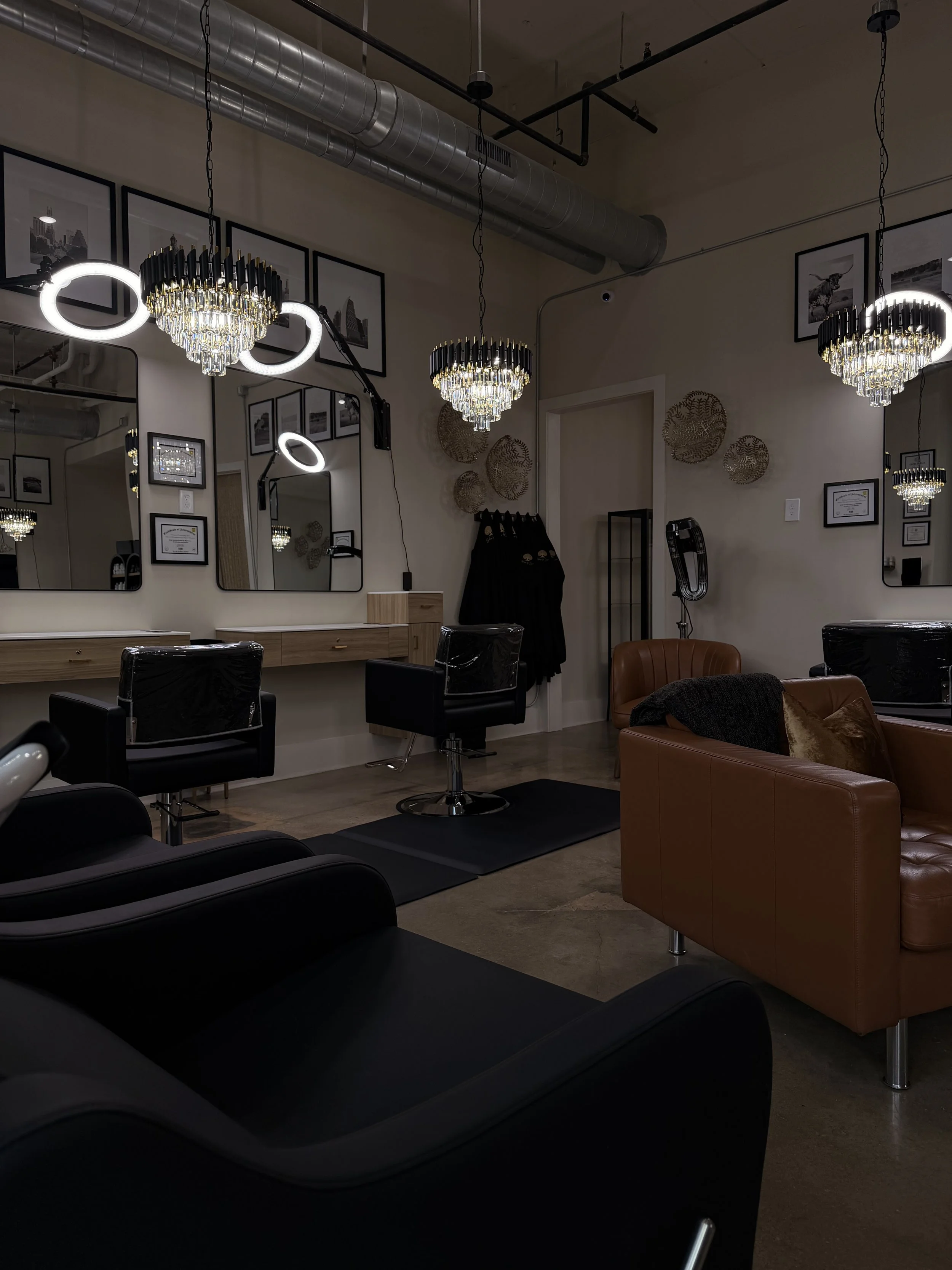 Interior of a modern hair salon with black chairs, a brown sofa, decorative mirrors, framed pictures, and elegant chandeliers with crystal accents.
