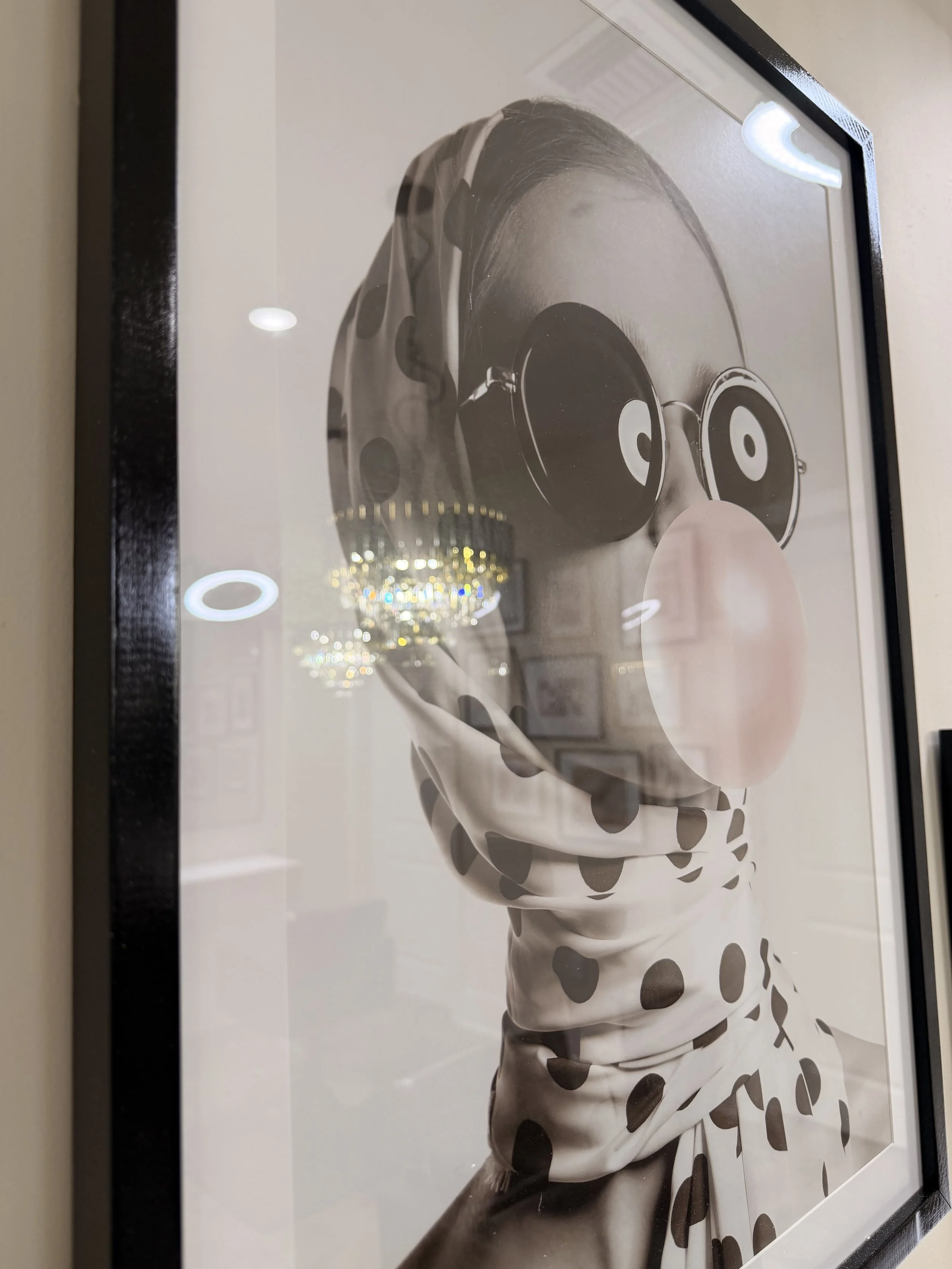 Black and white photograph of a woman with a bubblegum bubble and large round sunglasses, wearing a polka dot scarf, holding a camera up to her face.