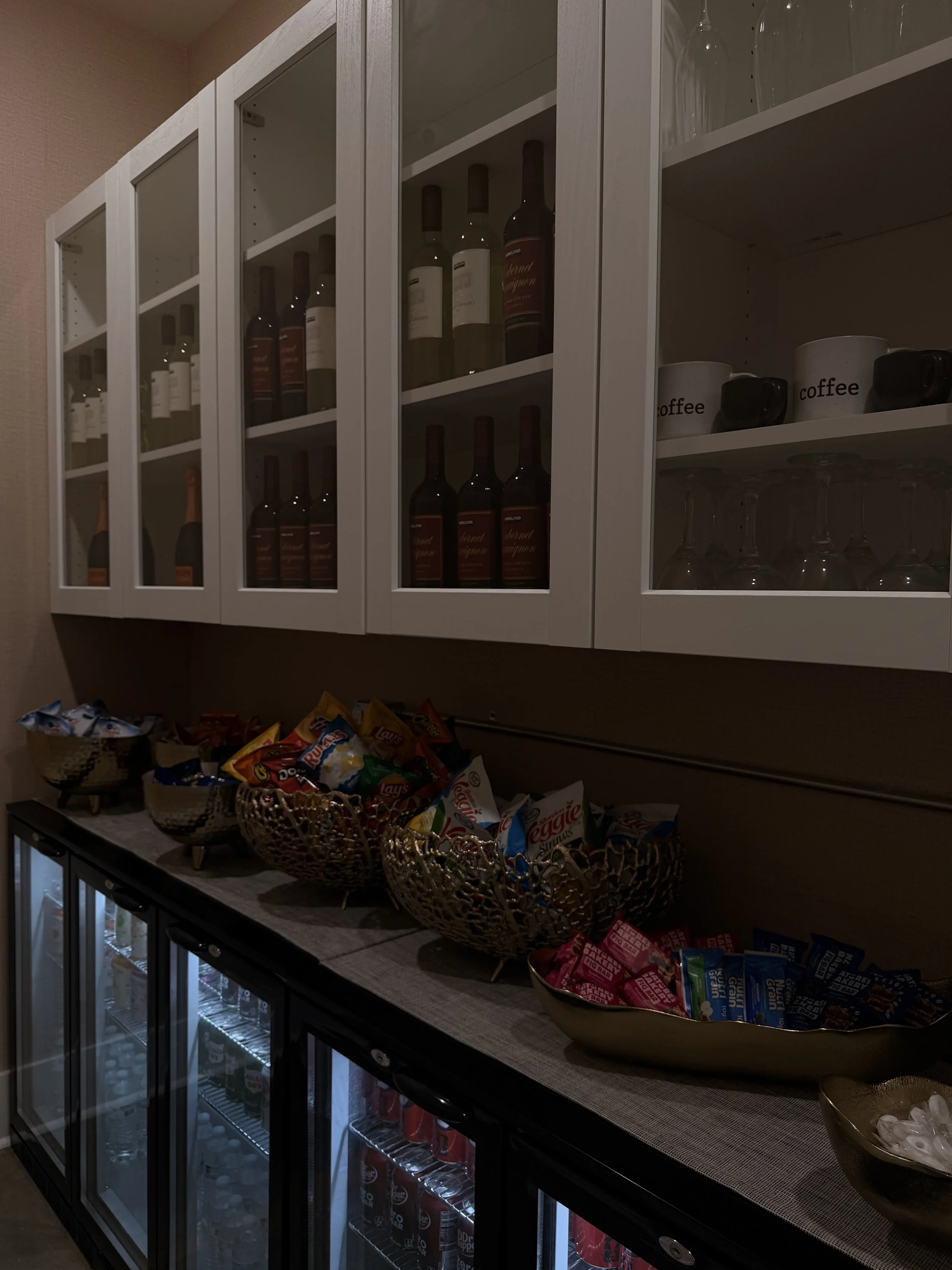 A snack and beverage station with glass-fronted wine cabinets, a countertop with bowls of snack packets, and a fridge beneath stocked with bottled drinks.