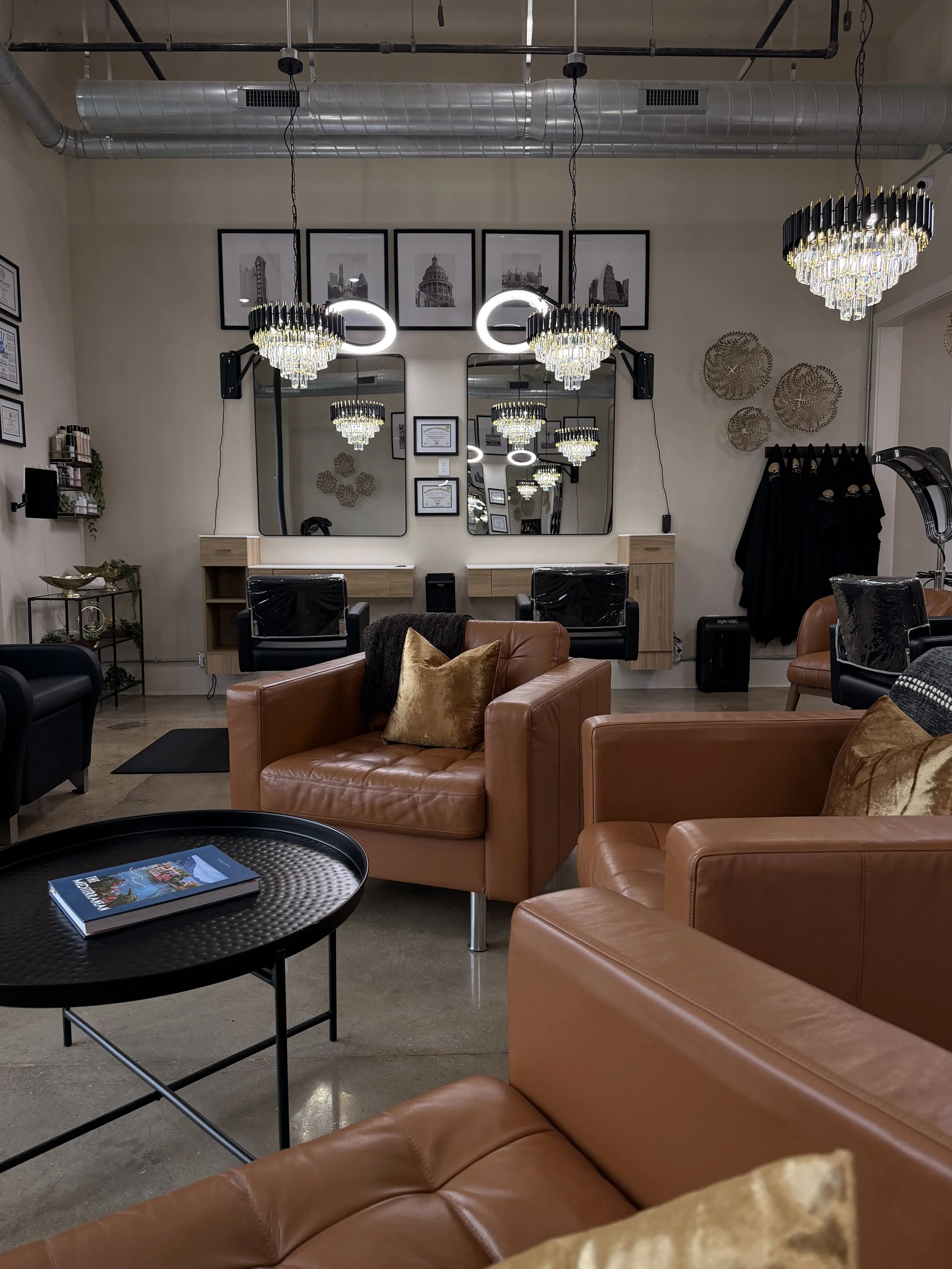 A modern salon room with leather armchairs, decorative pillows, and a black table with a magazine, mirrored walls, chandeliers, framed photographs, and certificates on the wall.