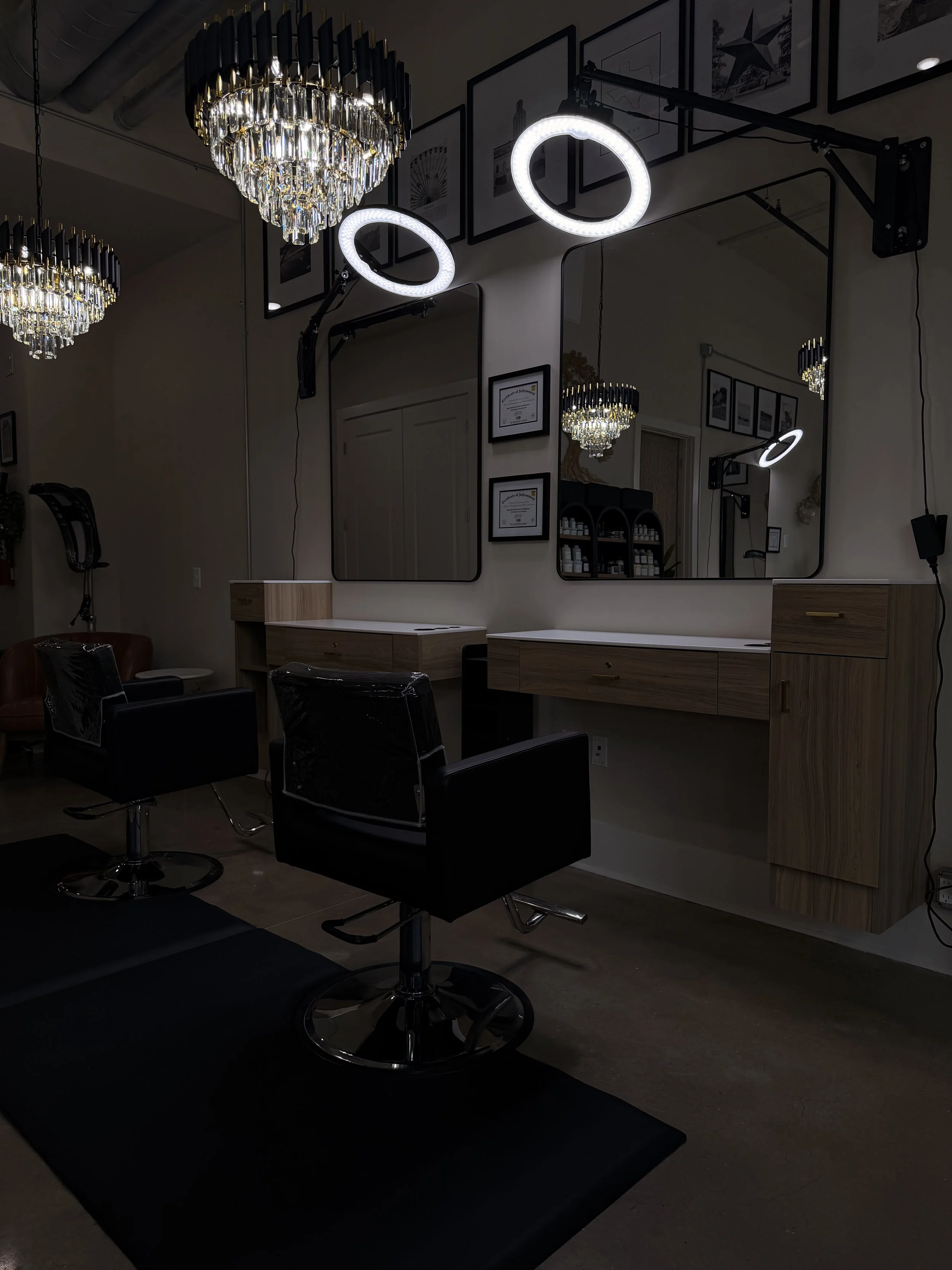 Salon with two black styling chairs in front of a large mirror with circular LED lights, chandeliers hanging from the ceiling, framed certificates and artwork on the walls, and wooden cabinets on the wall.