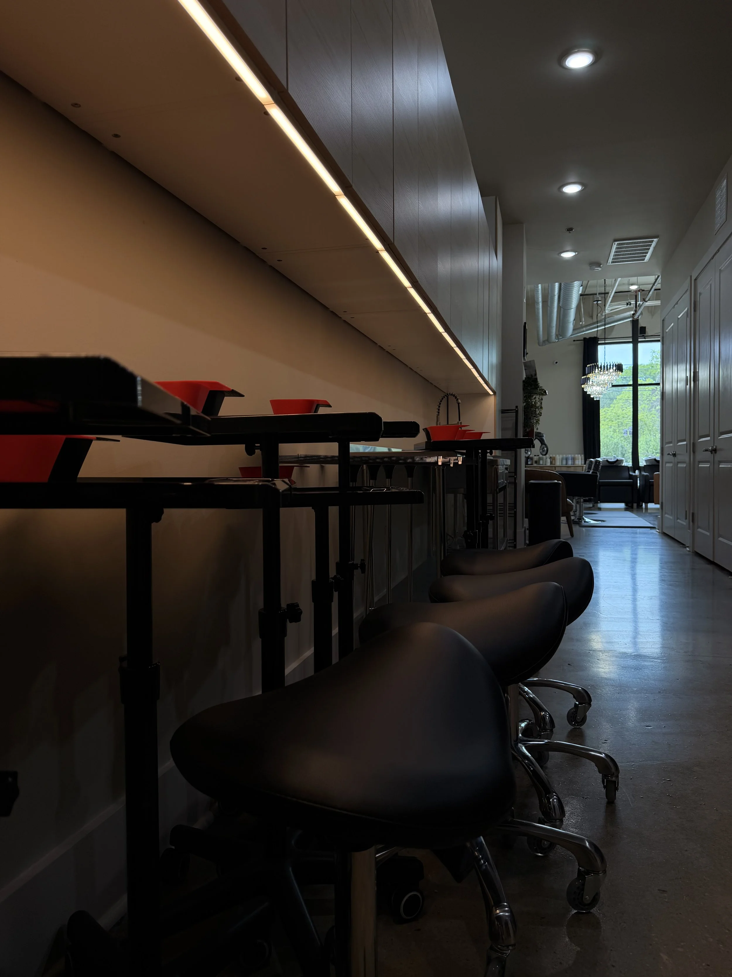 Empty bar-height workstations with black chairs and red bowls in a dimly lit modern lounge or cafe with a view of a bright room with large windows and a chandelier.