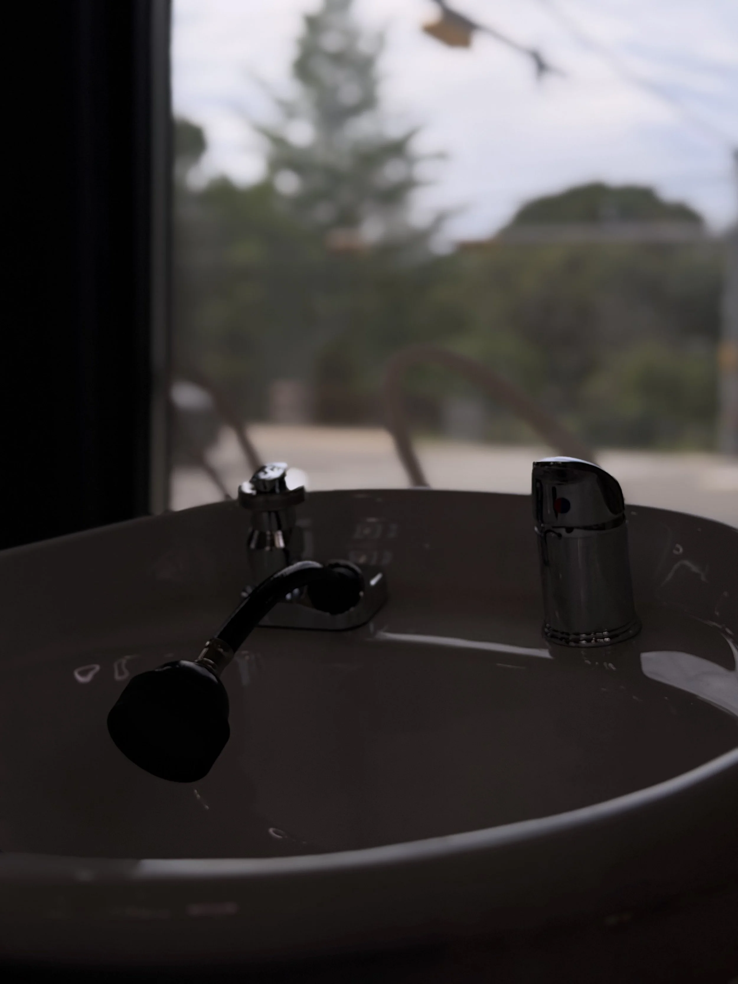 Close-up of a black facial steamer device on a white sink with a window in the background showing trees and a cloudy sky.