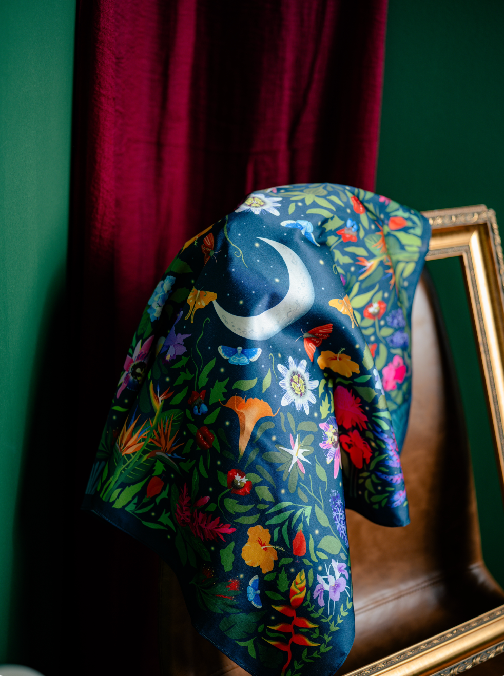 A colorful silk scarf with a nature and moon pattern draped over a brown leather chair, against green walls with red curtains and a gold picture frame.