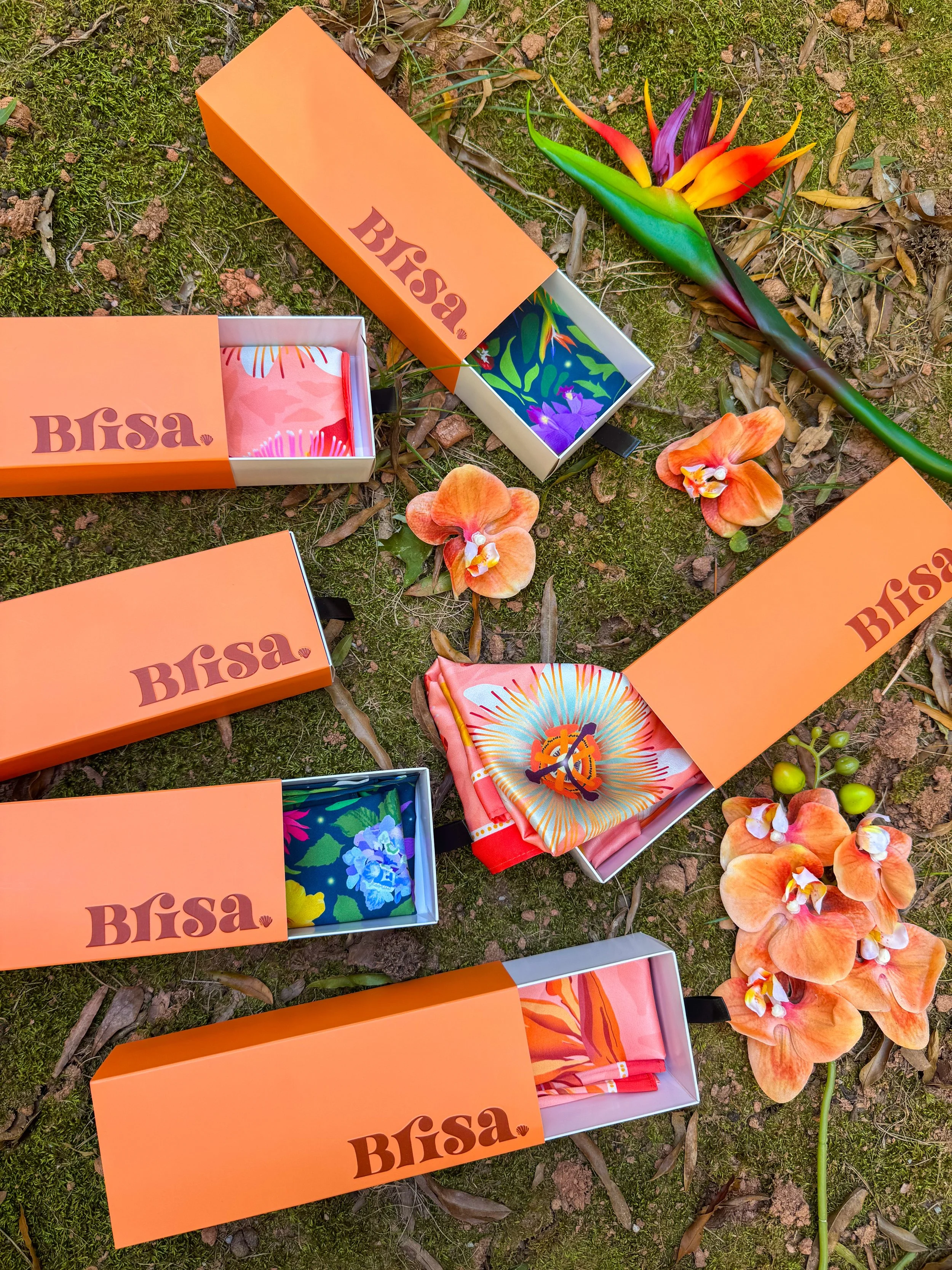 Multiple designer scarves displayed in open orange boxes labeled 'Blisa' on a mossy ground with orange orchids and a large tropical flower.
