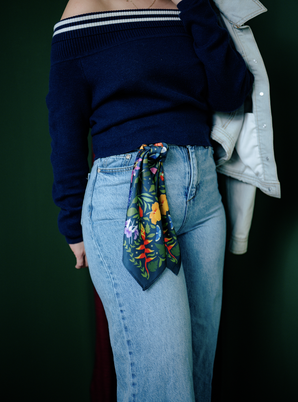 Close-up of a person wearing a navy off-the-shoulder sweater, light blue jeans with a colorful floral scarf tied at the waist, holding a light-colored denim jacket against a dark green background.