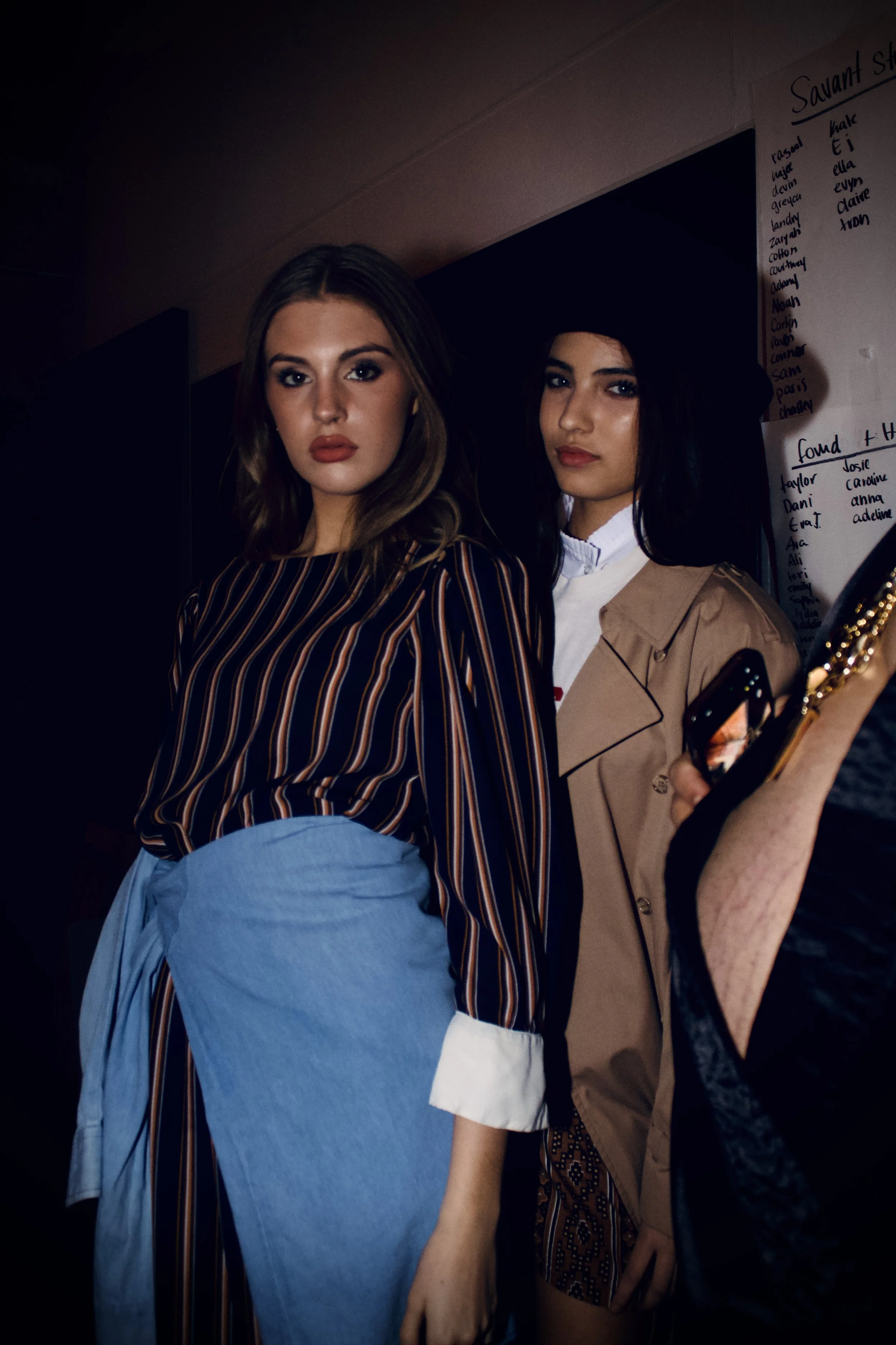 Two young women standing close together, one with shoulder-length light brown hair in a striped shirt and blue skirt, the other with long black hair wearing a tan coat and white shirt, in a dimly lit room with a whiteboard behind them.