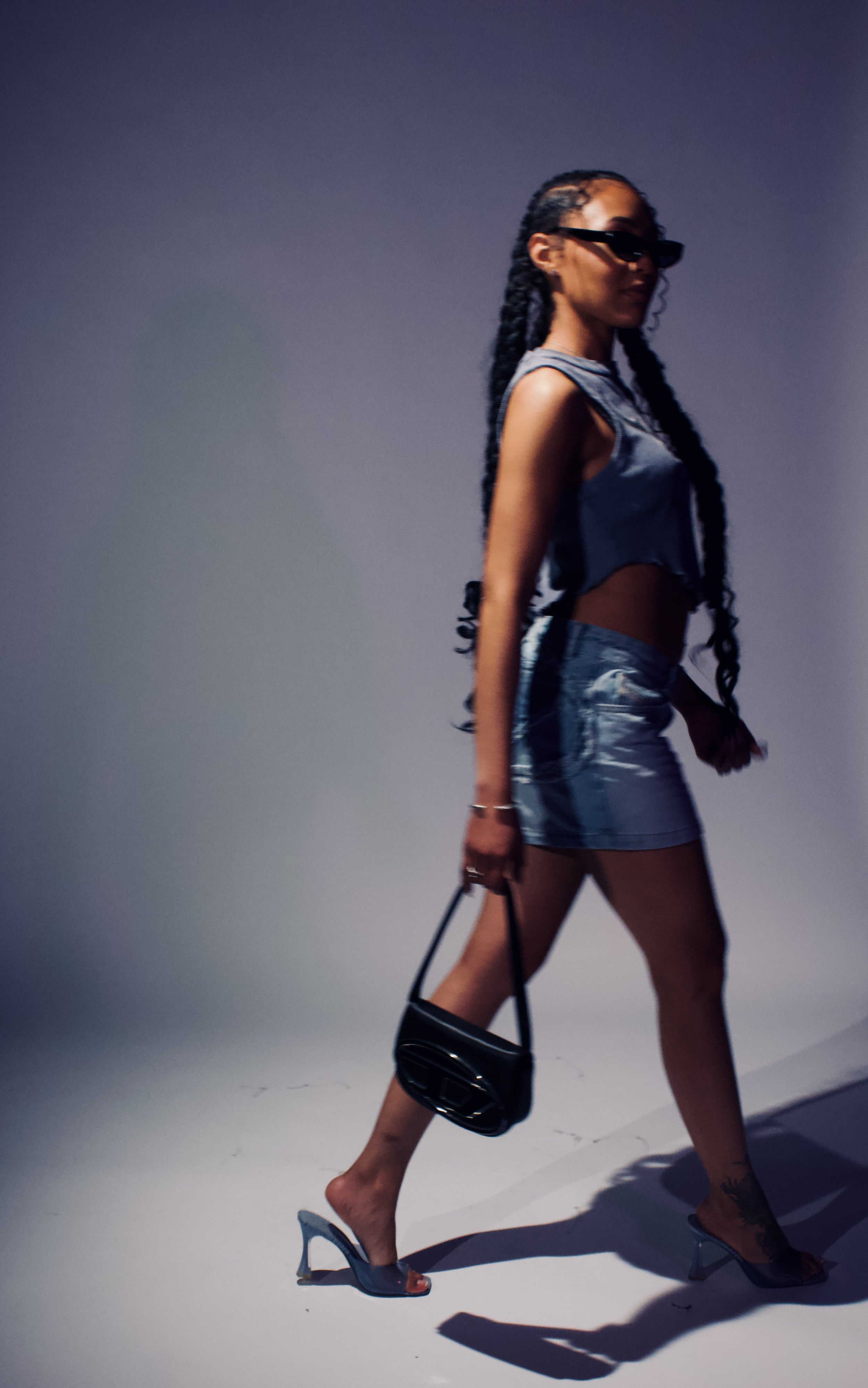 A woman with long braided hair, sunglasses, a sleeveless denim top, and a matching mini skirt, carrying a black purse and wearing high-heeled sandals, walking against a plain background.