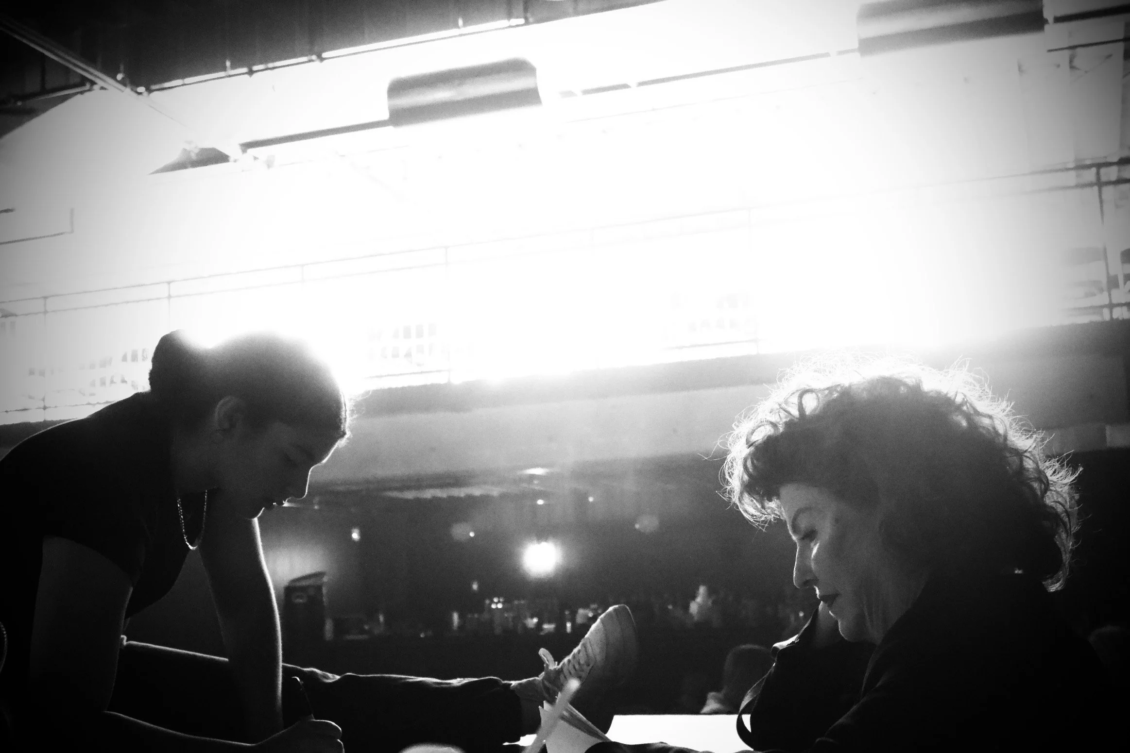 Two women sitting at a table, one writing and the other reading, with bright sunlight shining behind them.