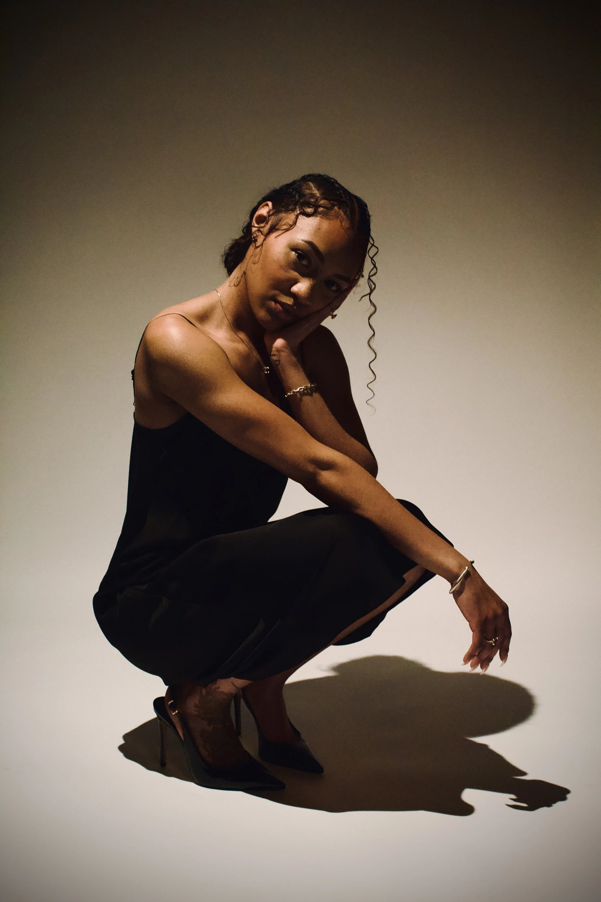A woman with curly hair posing in black high heels, crouching on the floor with her hand near her face, cast a shadow on the floor.