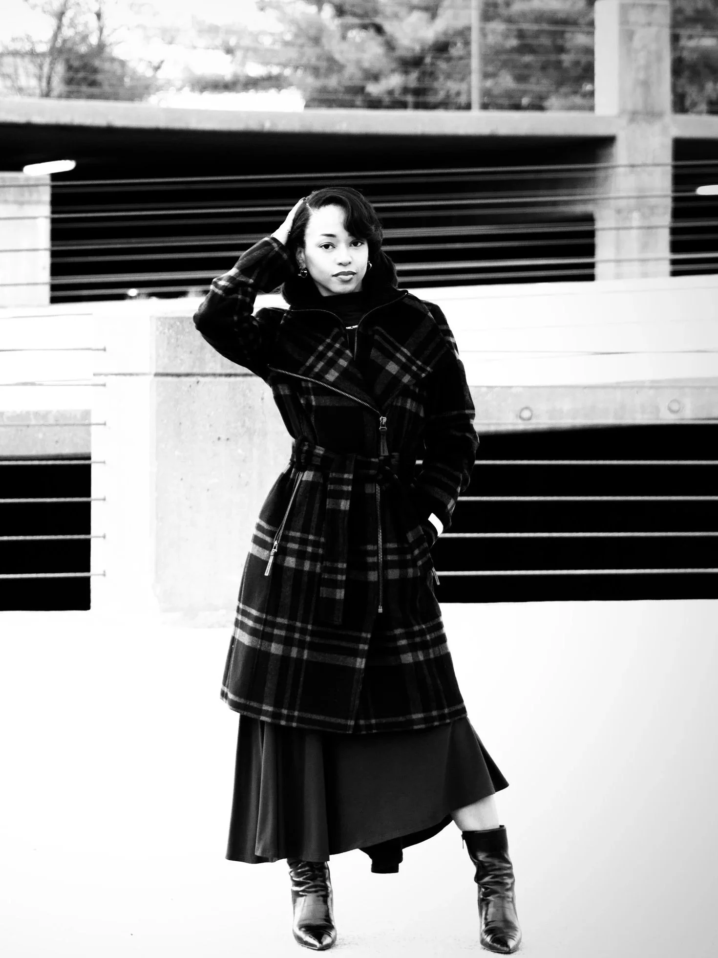A young woman standing outdoors in front of a concrete structure with horizontal railings, wearing a plaid coat, a long skirt, and heeled boots, looking at the camera.