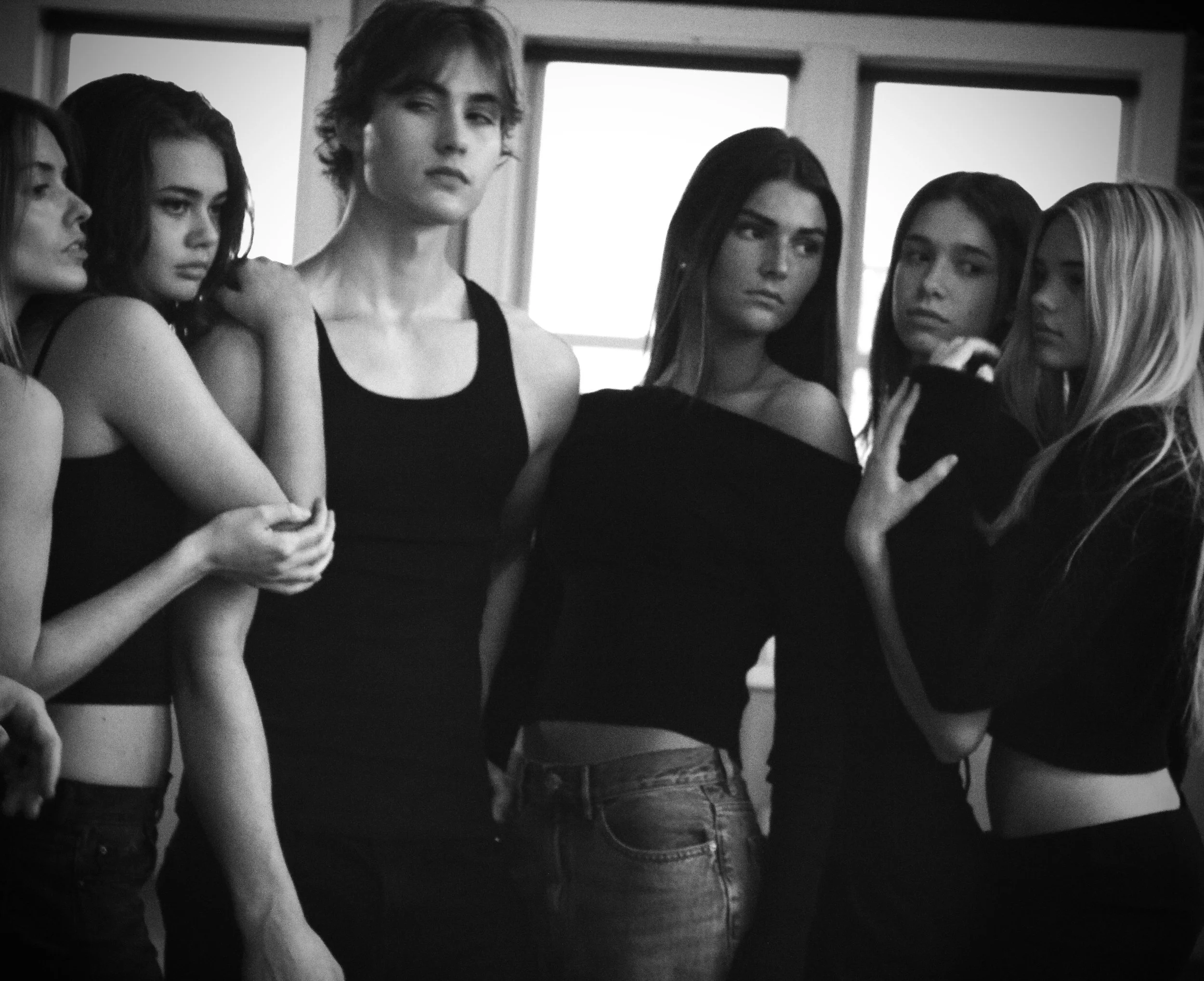 Black and white photo of six young women standing closely together indoors, some with arms around each other, looking serious or contemplative.
