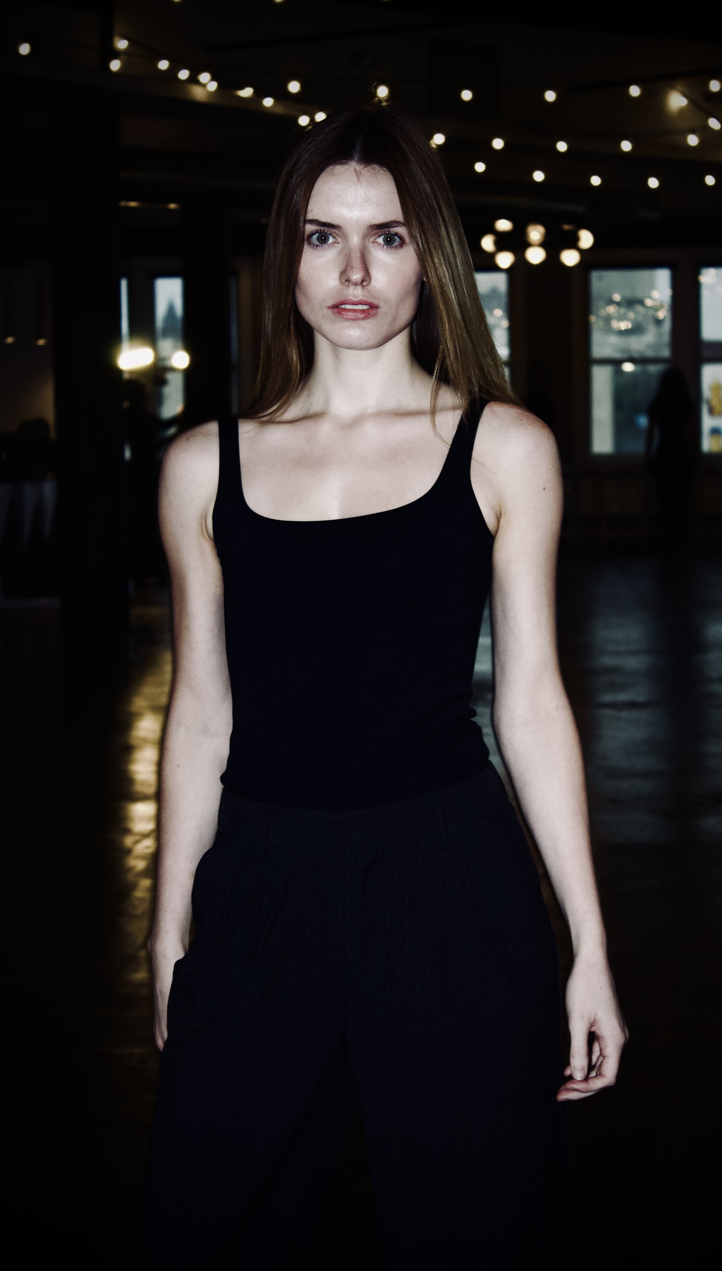 A young woman with long brown hair wearing a black tank top and black pants standing indoors with dim lighting and hanging ceiling lights.