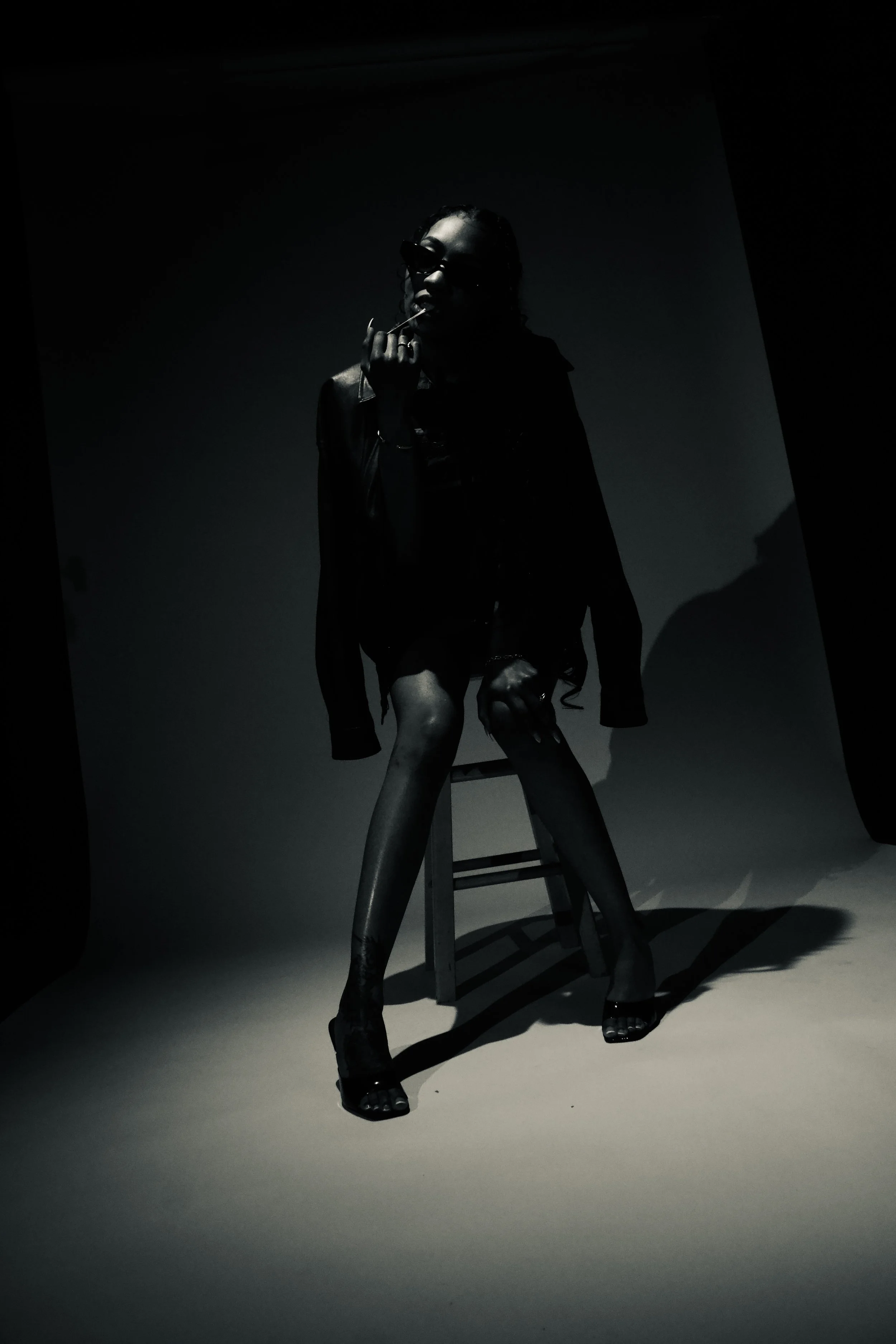 Darkly lit photo of a woman wearing sunglasses, jacket, and high heels, standing with one knee on a stool against a plain background.