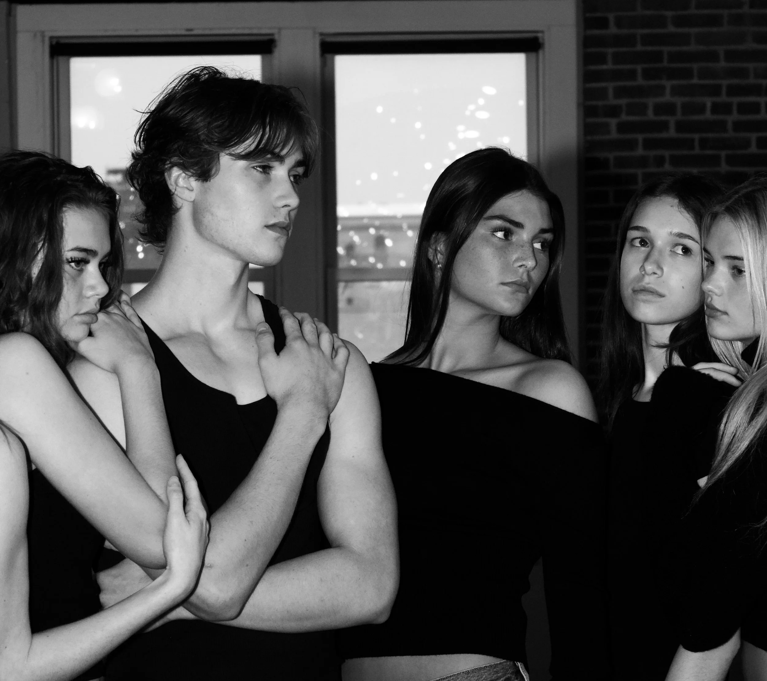 Five young people, four women and one man, standing close together indoors with a window behind them, displaying serious expressions.