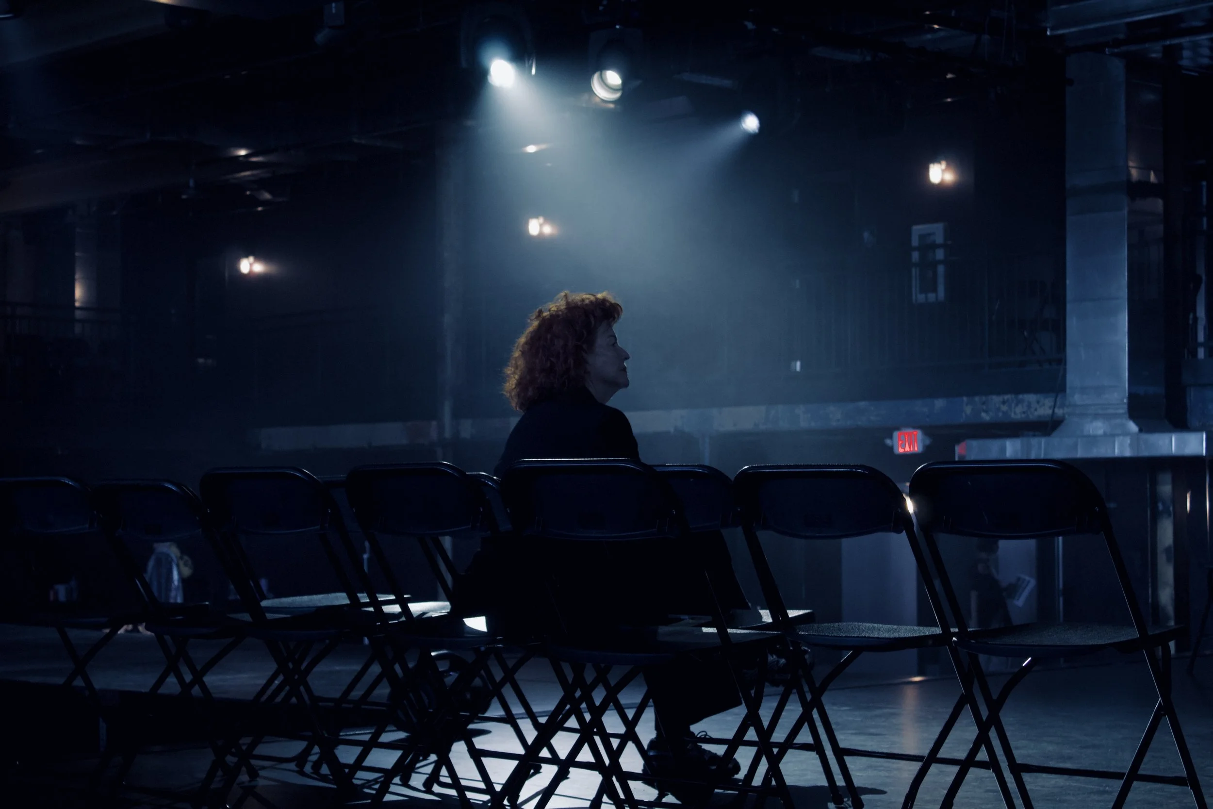 A woman sitting alone in a dimly lit auditorium, facing stage lights with empty chairs around her.