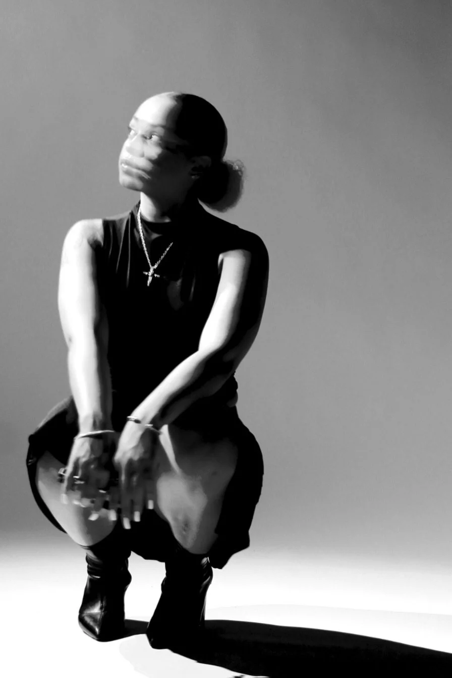 Black and white photo of a woman squatting with her head turned to the left, wearing a sleeveless top, necklaces, rings, bracelets, and boots, with a plain backdrop.
