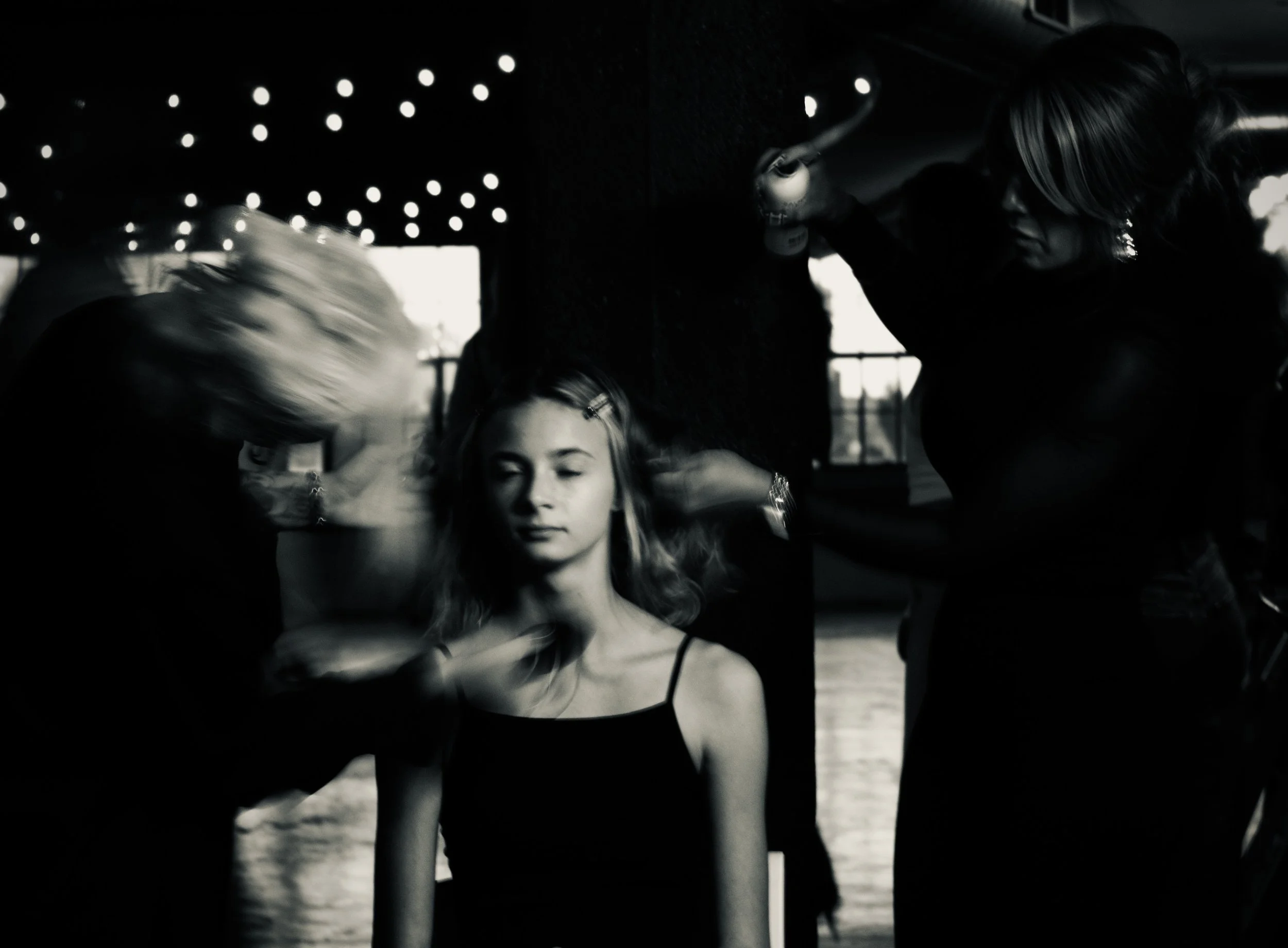 Black and white photo of a woman with closed eyes, getting her hair and makeup done by two stylists in a dimly lit space with string lights overhead.