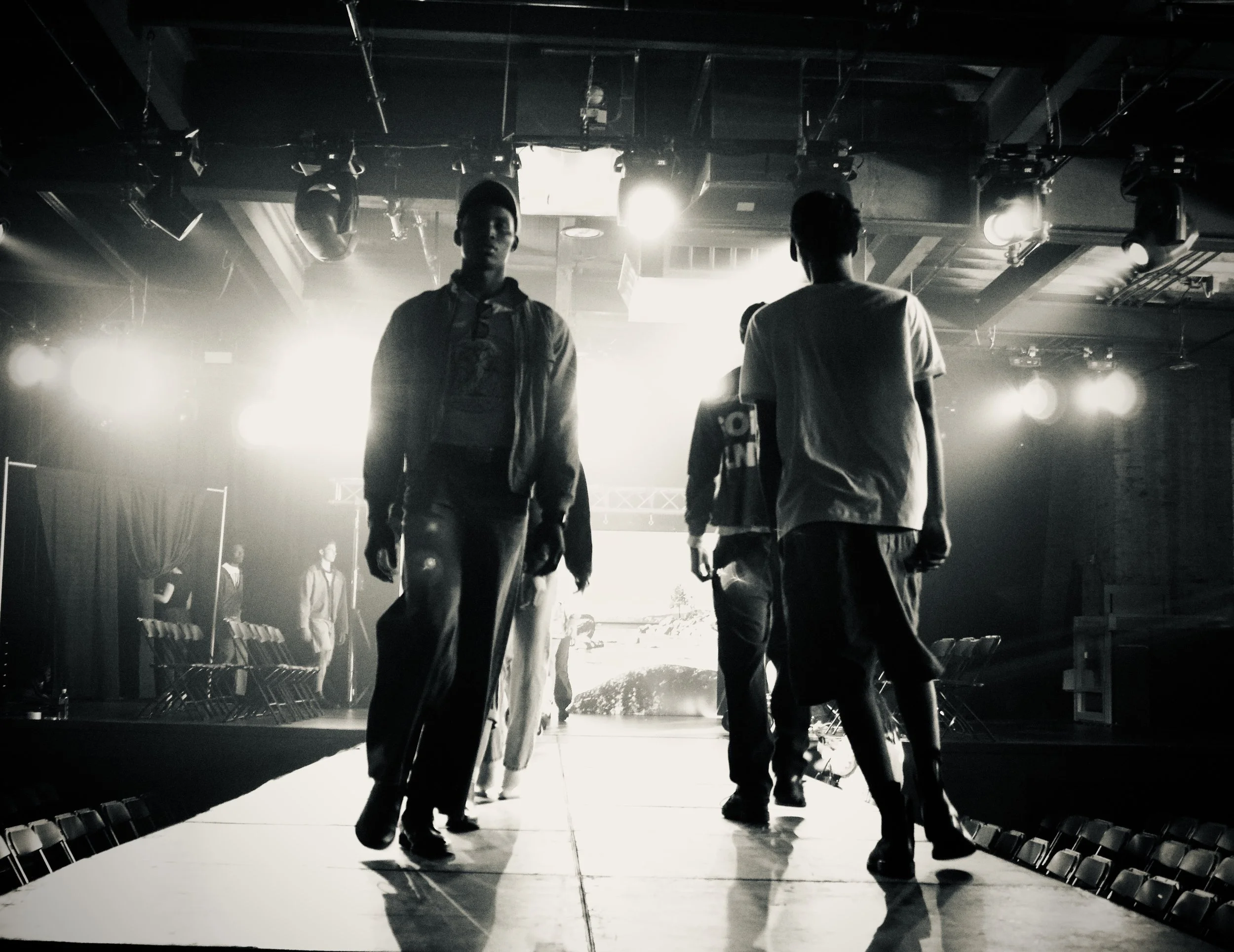 People walking on a runway in a dimly lit backstage area, with bright lights overhead and a large screen in the background.