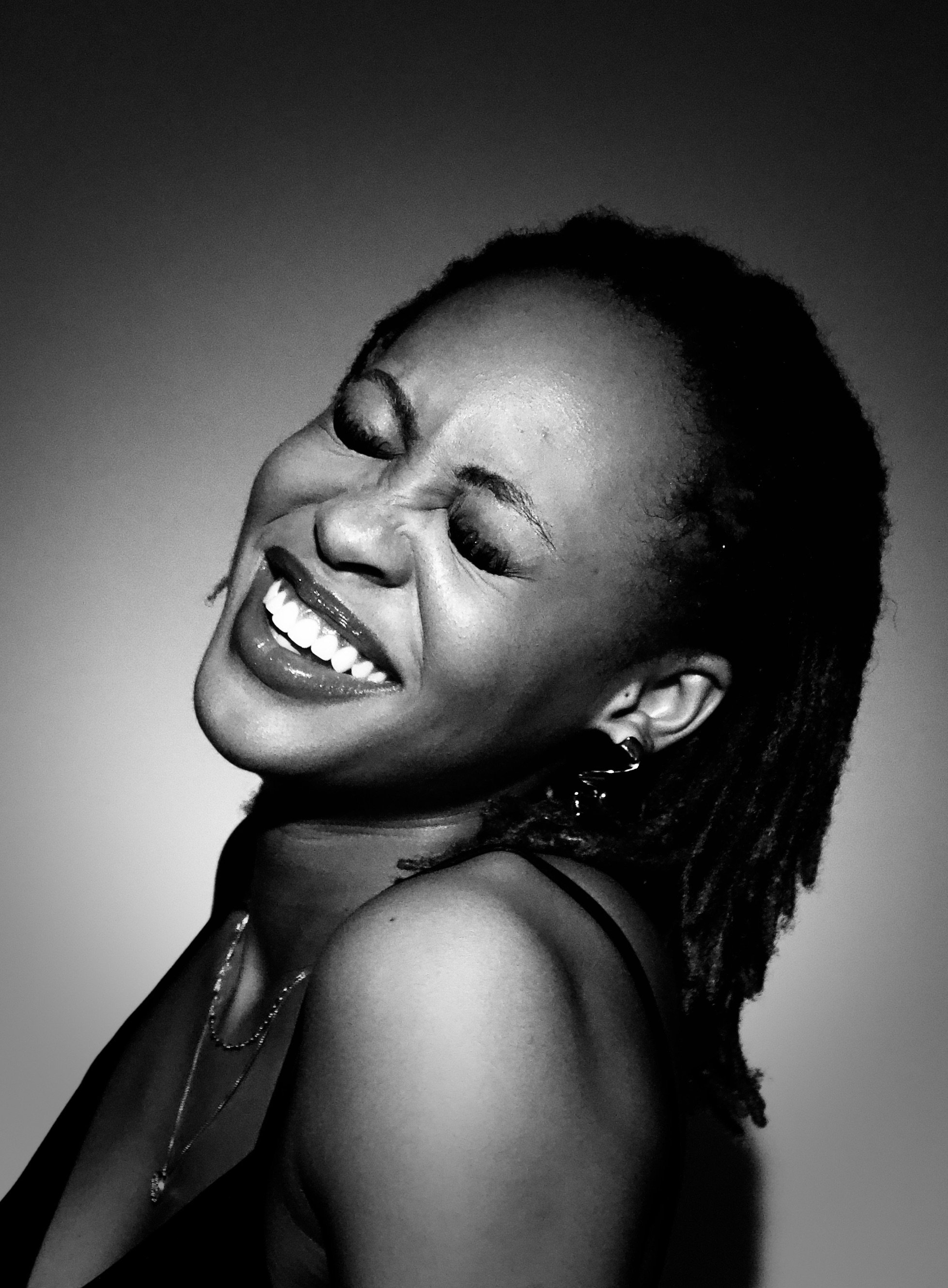 A woman with short dreadlocks smiling joyfully with eyes closed, in black and white, showing teeth and wearing earrings and a necklace.