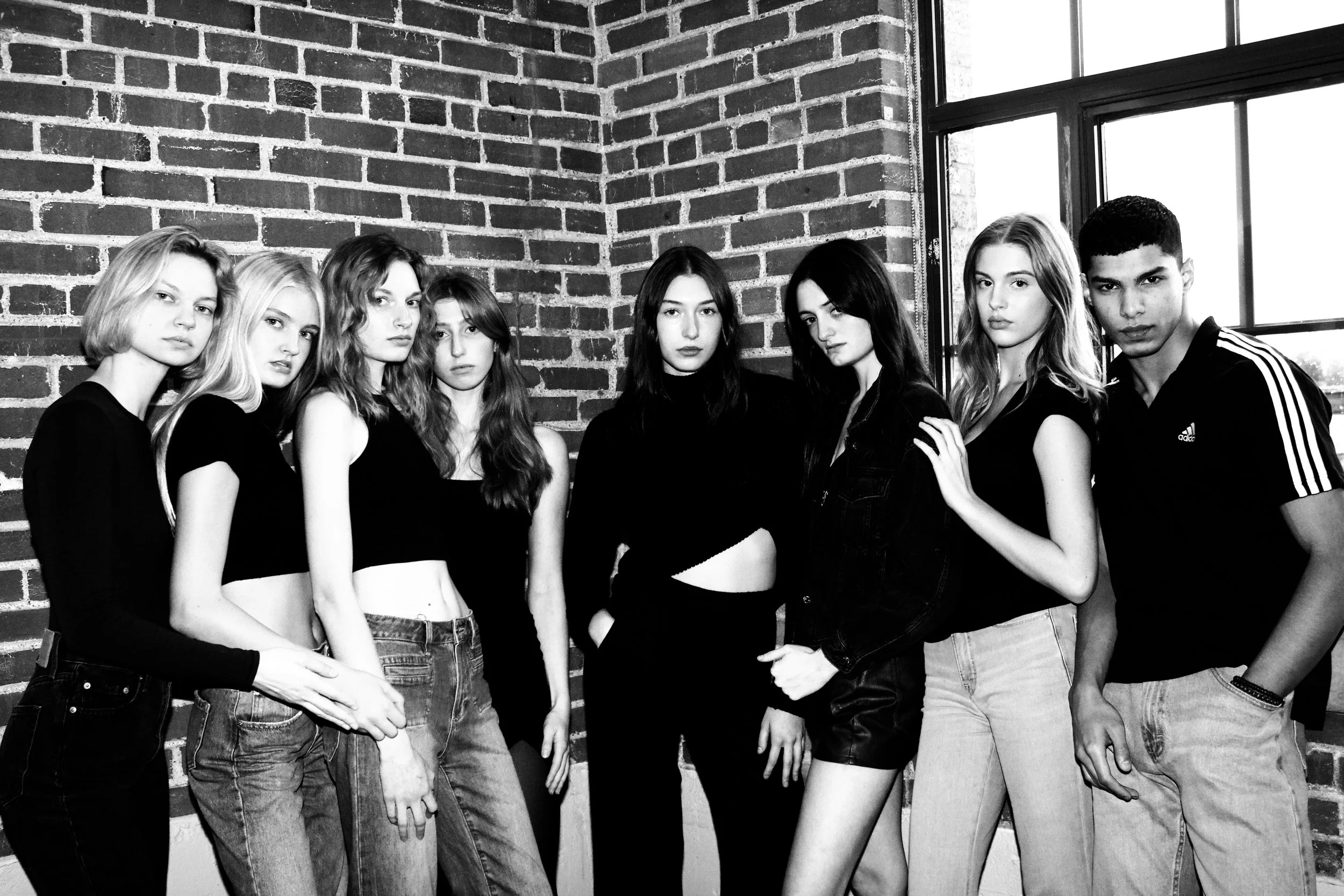 A group of eight young people, seven women and one man, standing together inside a room with a brick wall and a large window, all wearing black clothing.