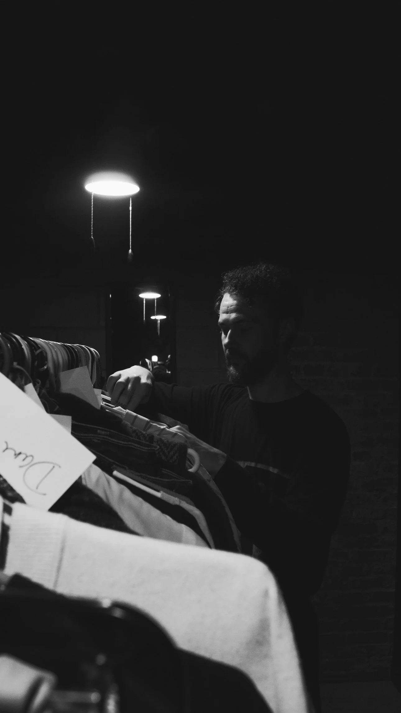 A man with a beard and curly hair looking at clothes on a rack in a dimly lit store, with ceiling lights reflected in a mirror.