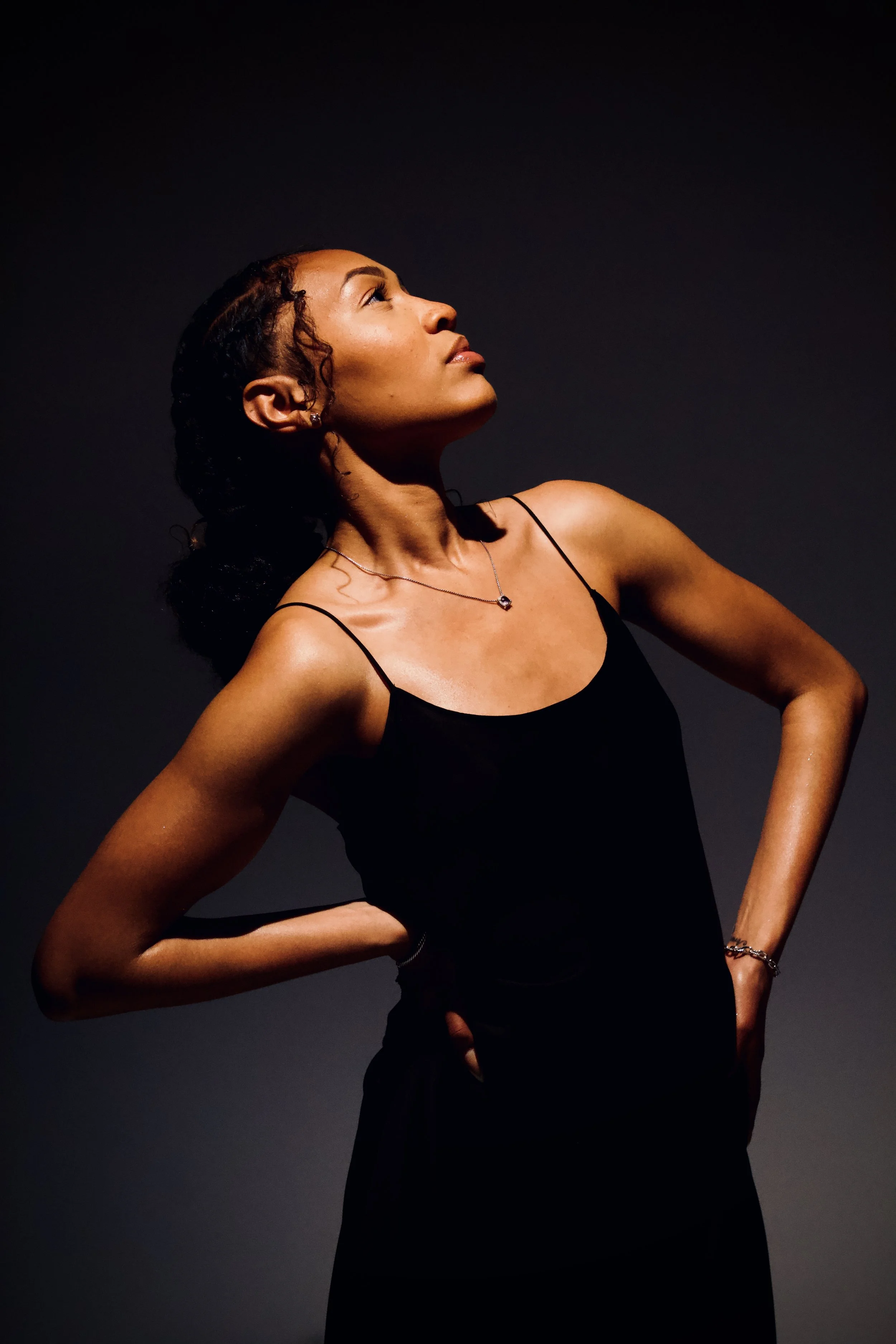 A woman with curly hair wearing a black dress and jewelry, standing against a dark background, looking upwards with her hands on her hips.