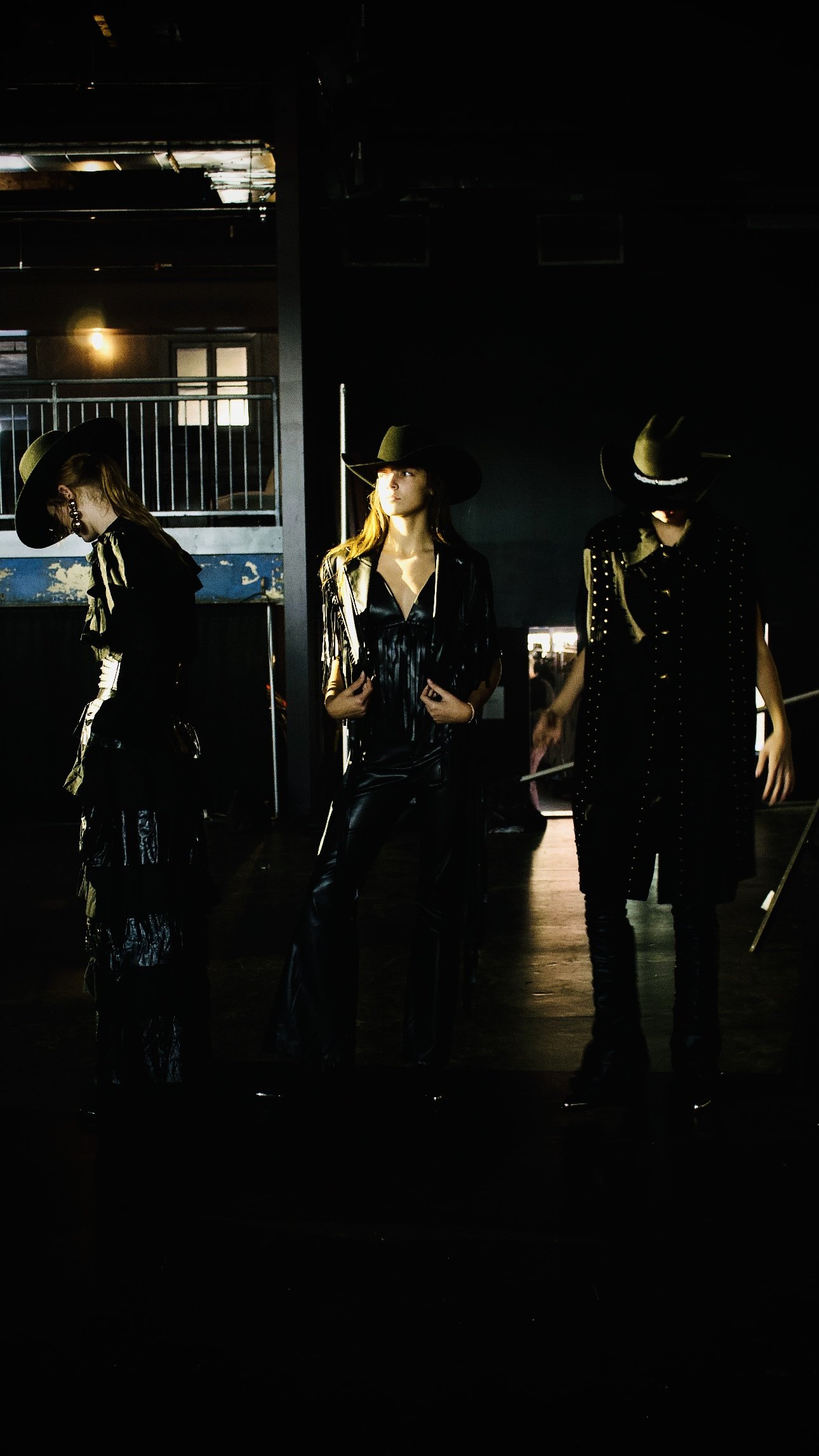 Three women dressed in black outfits with wide-brimmed hats, standing in a dimly lit indoor space.