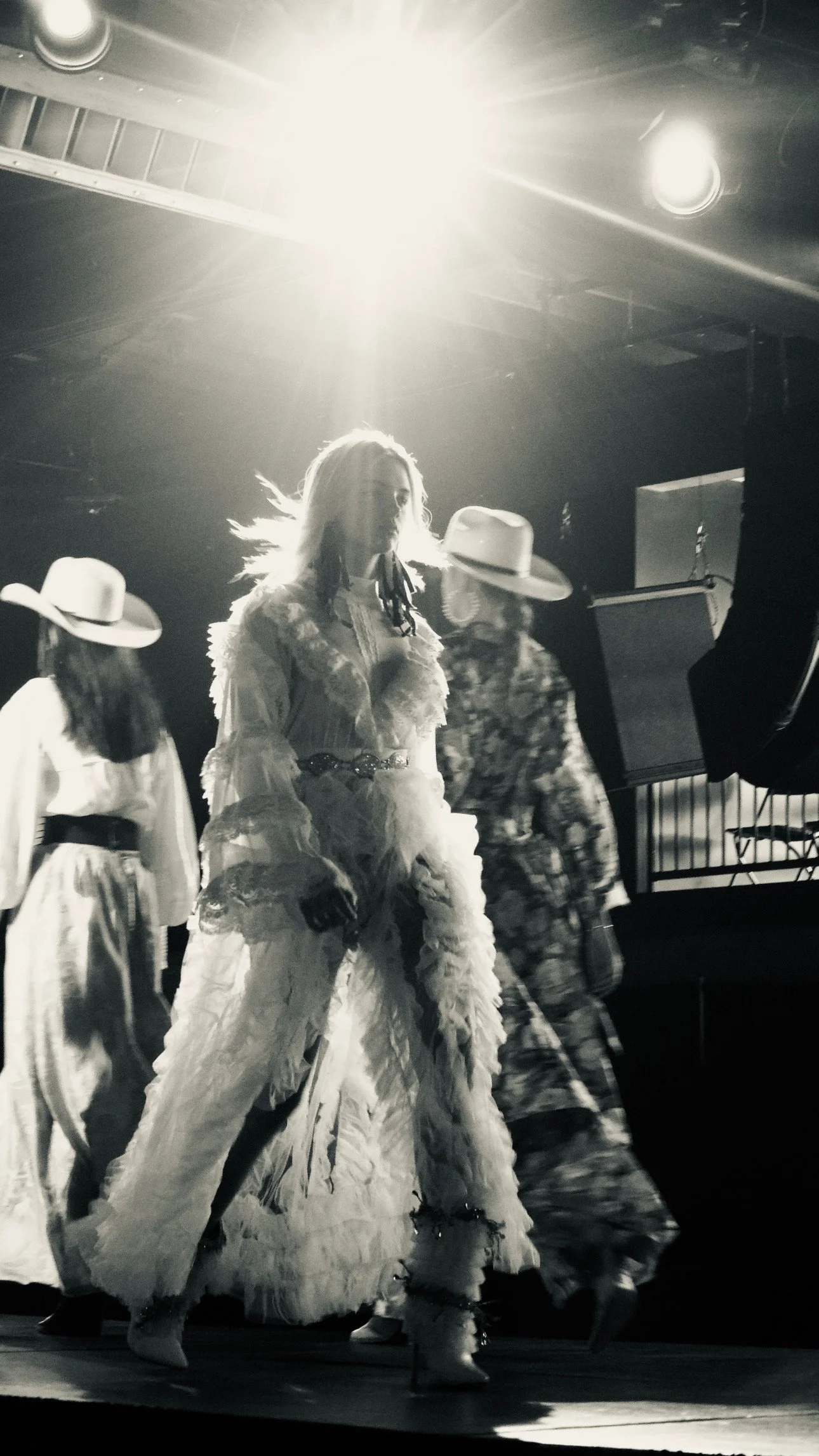 A black and white photo of a performer in vintage-style clothing on stage, backlit by a bright light. Other performers in similar vintage outfits and wide-brimmed hats are visible in the background.