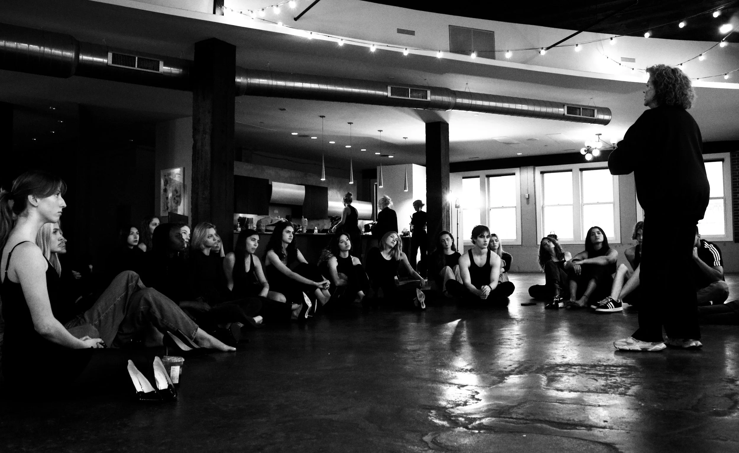 A group of young women sitting on the floor in a dance studio, attentively listening to a woman standing and speaking to them. The studio has large windows, string lights, and a modern interior with wood beams and hanging lights.