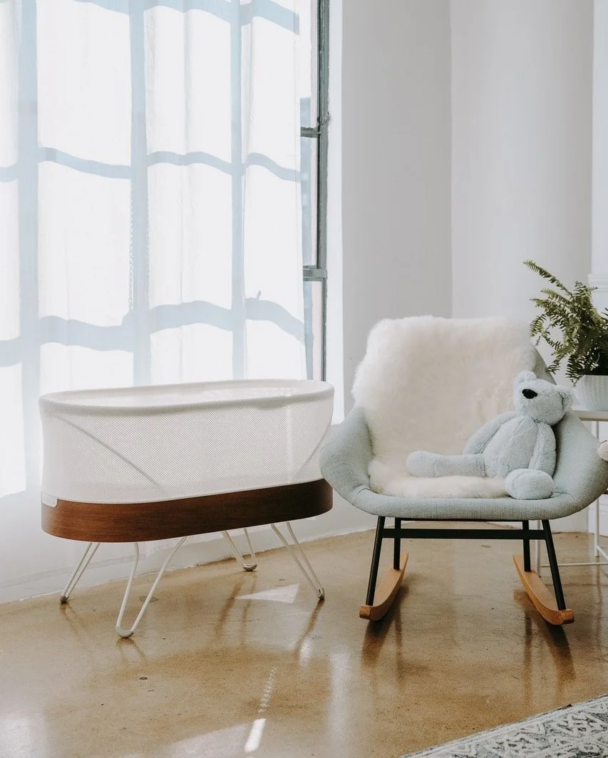 A cozy interior corner with white curtains, a modern white and wood rocking cradle, a light gray rocking chair with a plush teddy bear, and a potted fern plant.