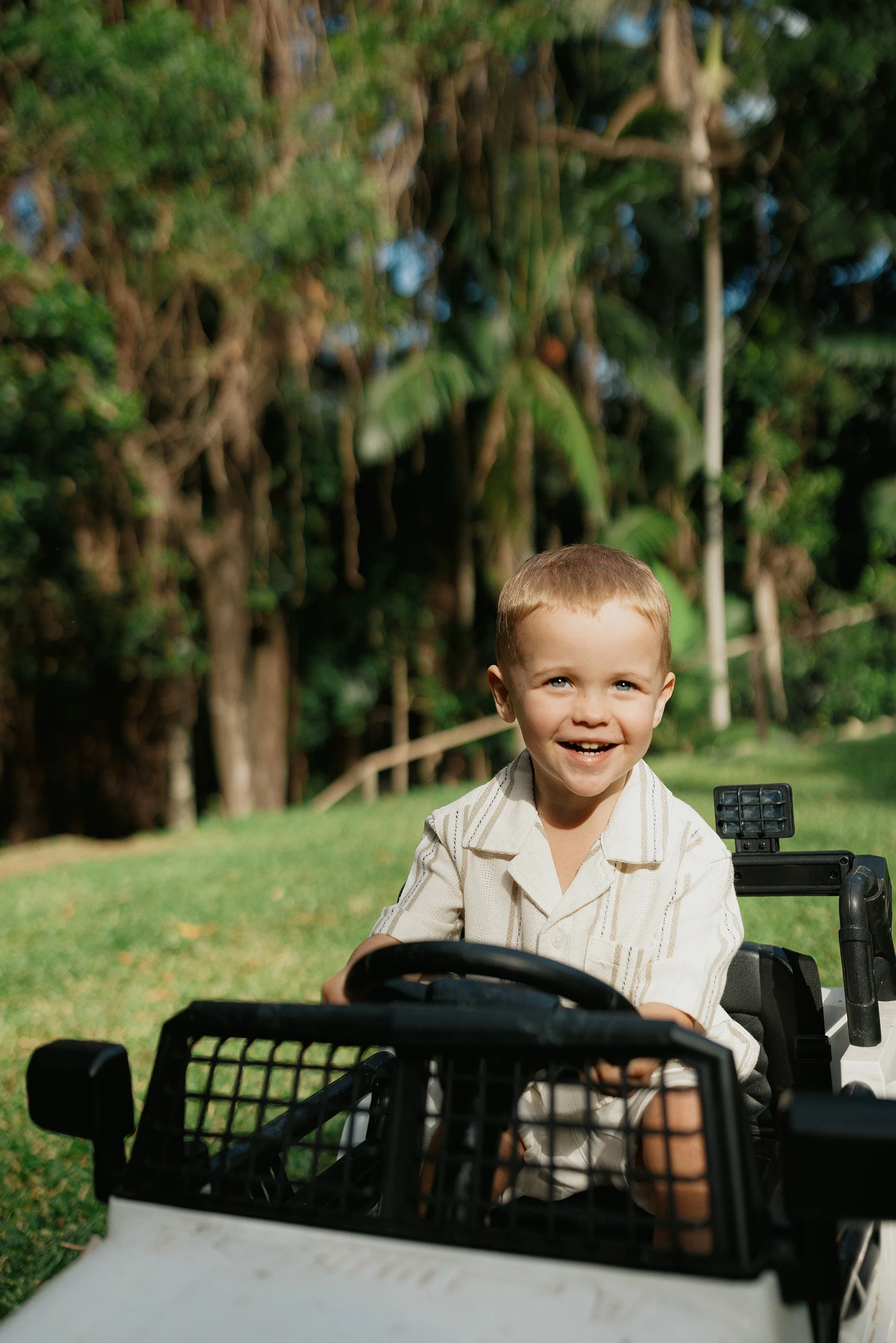 A young boy smiling and sitting in a toy car outdoors on a grassy area with trees in the background.