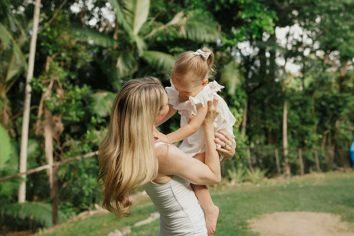A woman holding a young girl in her arms outdoors with green trees and bushes in the background