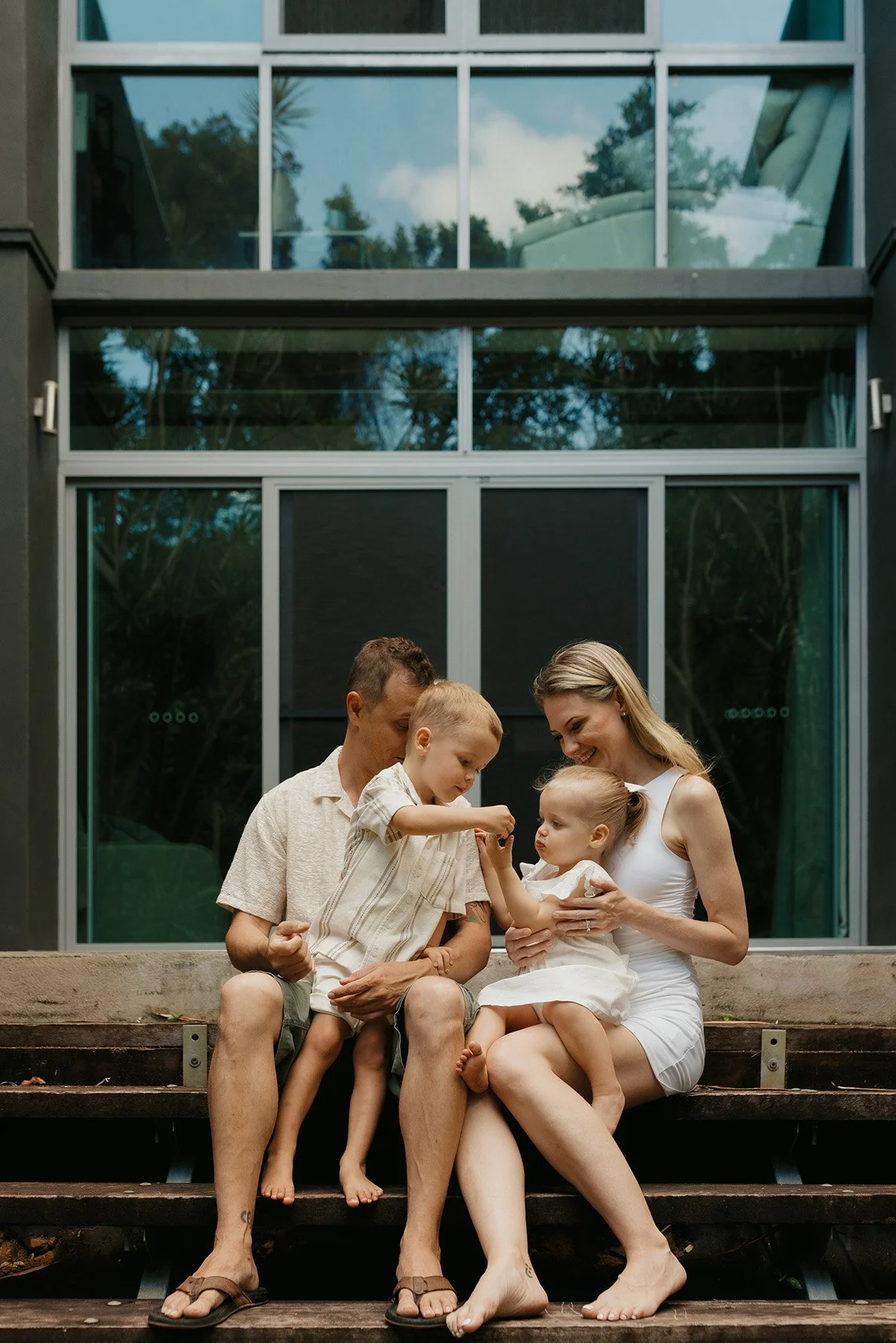 A happy family of four sitting on wooden steps outside a modern house with large glass windows. The mother holds a young daughter, and a father with two young boys is interacting with them, all smiling.